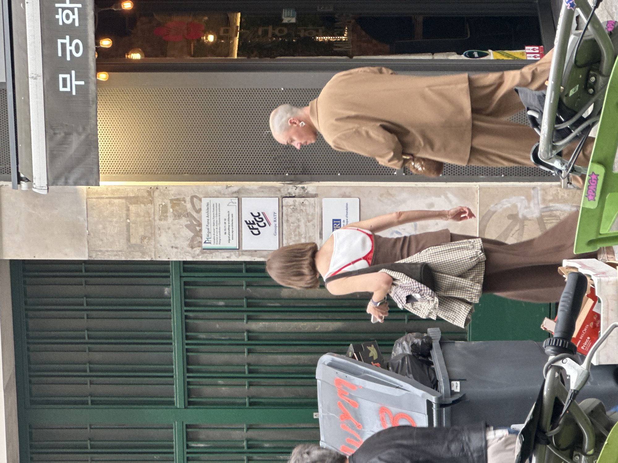 The image captures a street scene in Paris, France. A building facade serves as the backdrop, with signage in Korean script visible. Two pedestrians are in motion: one is wearing a brown suit, and another is clad in a white and brown top with brown pants. A grey trash bin is prominently placed in the foreground, partially obscuring another pedestrian in the bottom center. In front of the bin is a parked bicycle. Signage is attached to the wall. The scene includes elements of daily urban life.