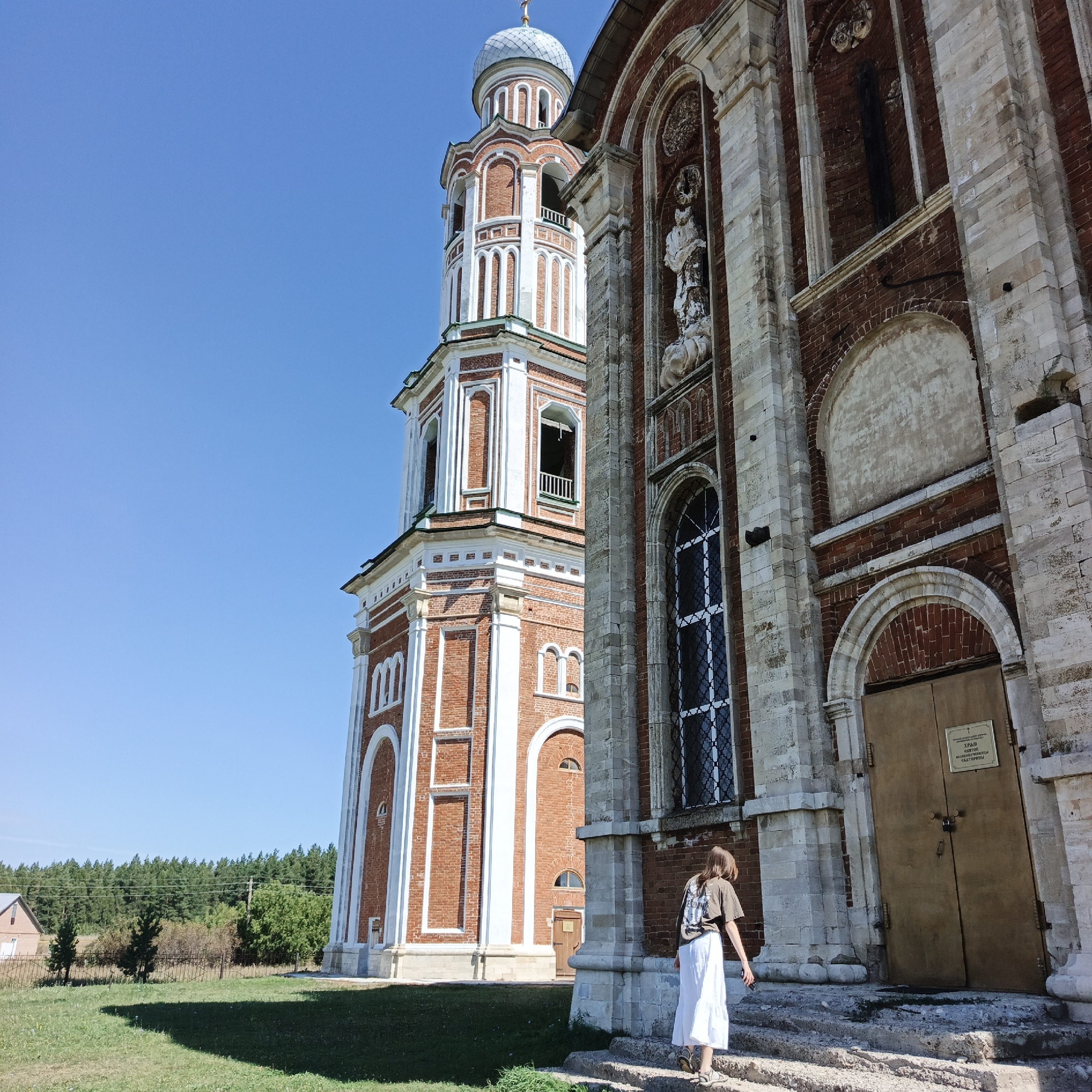 A wide-angle, eye-level outdoor shot captures a church tower and a portion of the church facade on a clear, sunny day. A young woman with long brown hair, wearing a dark t-shirt and a white skirt, is walking up a stone staircase towards the church entrance. She is carrying a phone in her right hand. The church is constructed of red brick with white decorative elements and stone accents. The tower, to the left, is tall and multi-tiered, topped with a silver dome. The church facade, to the right, features a large arched window with intricate white tracery and a large, plain brown double door with a sign above it. The church grounds consist of a grassy area in the foreground with a shadow cast by the building, and a line of trees in the background. A small house and a fence are visible to the far left. The scene is located in the village of Tugustemer, Russia, likely at the Church of St. Nicholas.