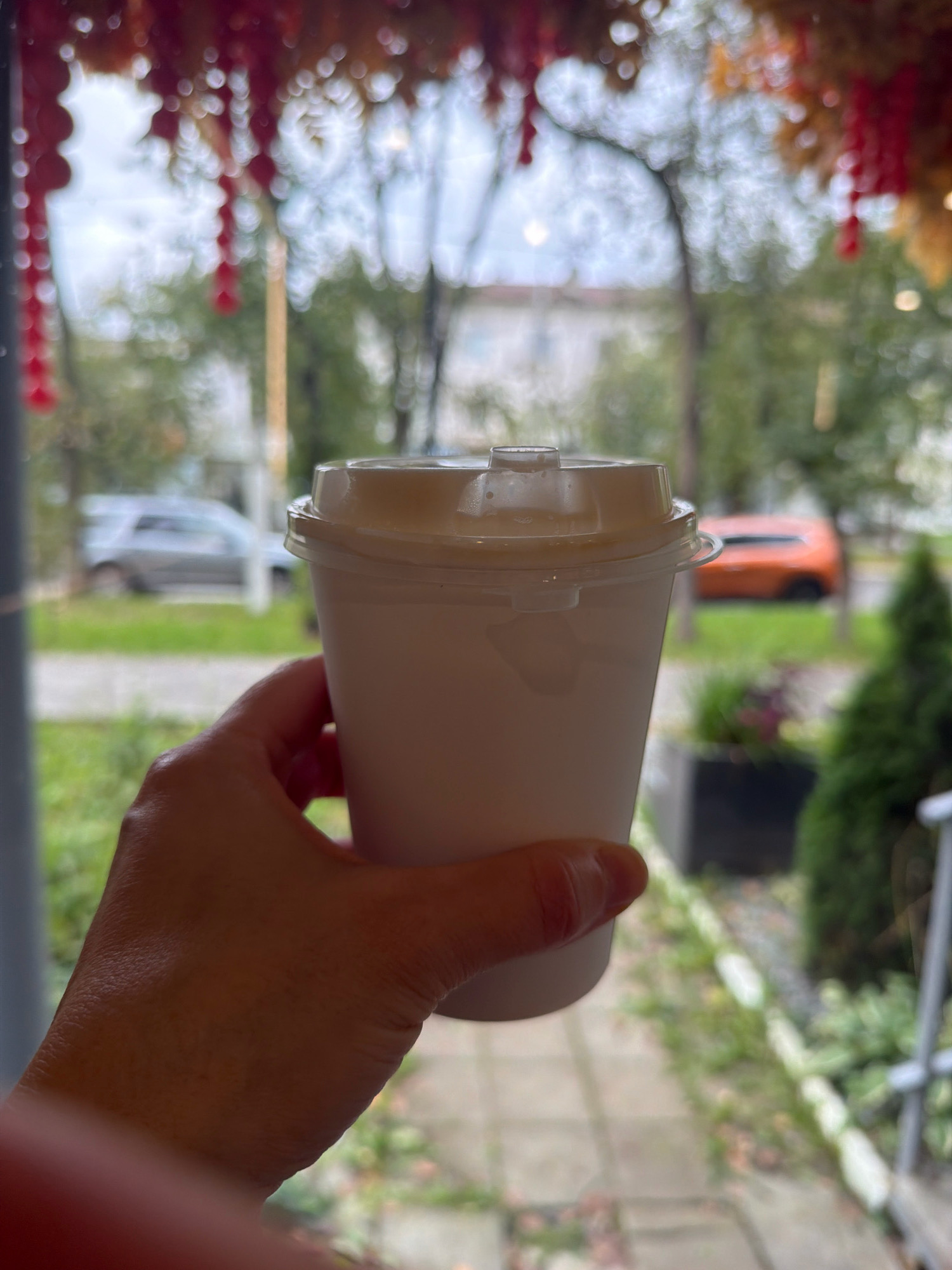 A hand holds a white disposable coffee cup with a clear plastic lid, positioned in the foreground. The cup is out of focus. In the background, viewed through a window, is an outdoor scene. There is a paved walkway and a grassy area with some foliage. To the left, a silver SUV is parked on the side of a street. Further to the right, an orange car is visible. The sky is overcast. Red and yellow decorative elements hang from the top of the frame, obscuring part of the background and creating a bokeh effect. The scene is likely in an urban or suburban area, possibly a cafe or shop with a view of the street in Chaikovsky, Russia, based on the provided location context.