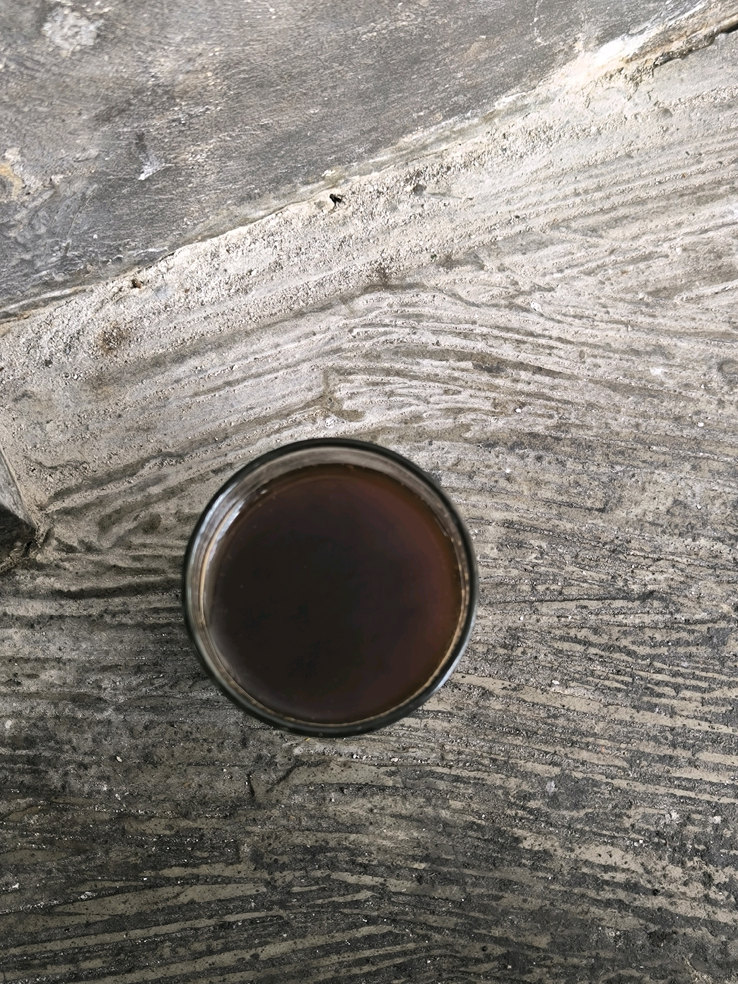A top-down shot shows a clear glass filled with a dark liquid, likely coffee or tea, sitting on a textured, grey, stone-like surface. The surface exhibits distinct striations and appears weathered, with patches of lighter material embedded within the darker grey. The glass is positioned in the lower center of the frame, its rim clearly defined against the textured background. The lighting is neutral, casting no strong shadows, and the focus is sharp on both the glass and the surface it rests upon. The scene is static and appears to be outdoors, given the texture of the surface, possibly a patio or walkway. There are no people or animals visible in the image.