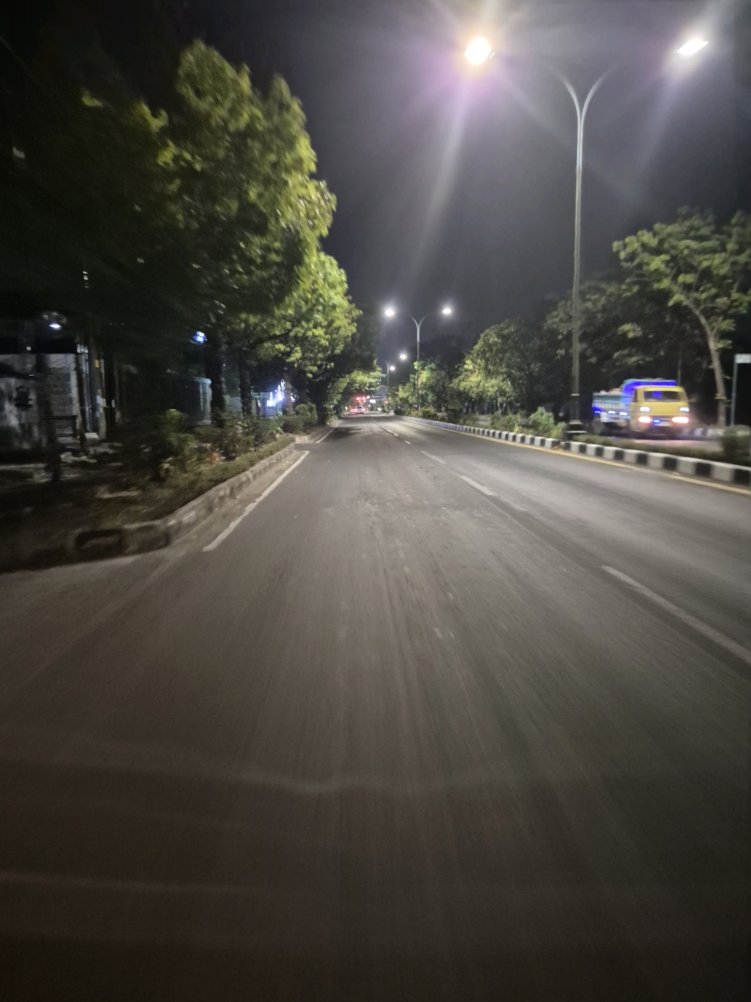 An eye-level, forward-facing, slightly blurry nighttime view of a paved road in Surakarta, Indonesia. The road appears to be a multi-lane street with white lane markers visible. On the right side of the road, a yellow and blue truck is parked. Trees line the right side of the road, with streetlights illuminating the scene. The left side of the road features more trees and some buildings, also dimly lit. The overall atmosphere is dark and quiet, suggesting a residential or commercial area at night.