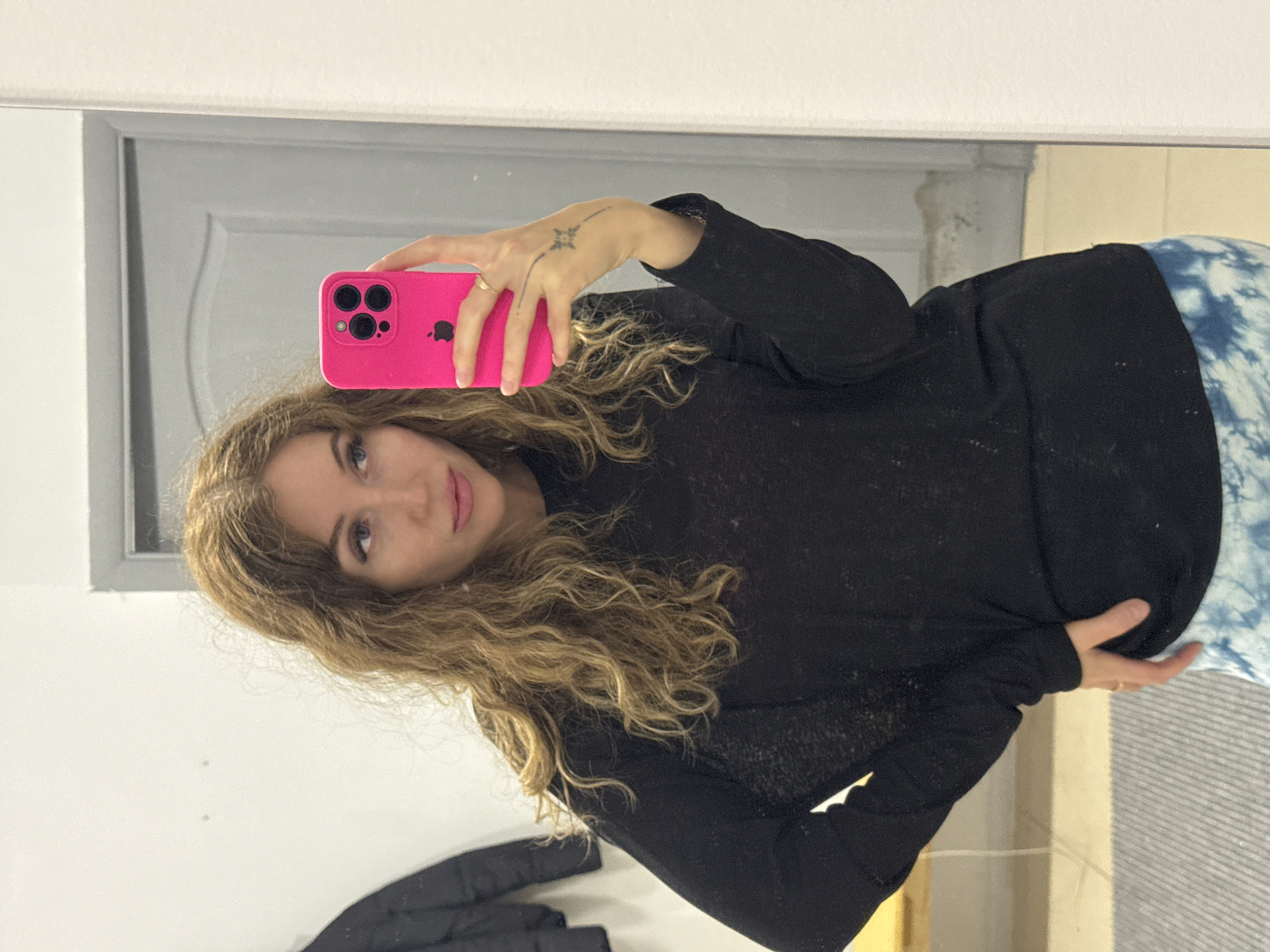 A mirror selfie captures a woman holding a hot pink iPhone with a visible Apple logo. The woman has long, wavy blonde or light brown hair and light-colored eyes, looking towards the camera with a soft expression and slight smile. She is wearing a black long-sleeved top, which appears to be a lightweight or sheer fabric. Her left hand, holding the phone, has a small tattoo on the back, resembling a star or compass design with a line. A ring is visible on her finger. Below the black top, a garment with a blue and white tie-dye pattern is visible, possibly pants or a skirt. The background, reflected in the mirror, shows a light-colored wall and a grey door or doorframe, indicating an indoor setting.