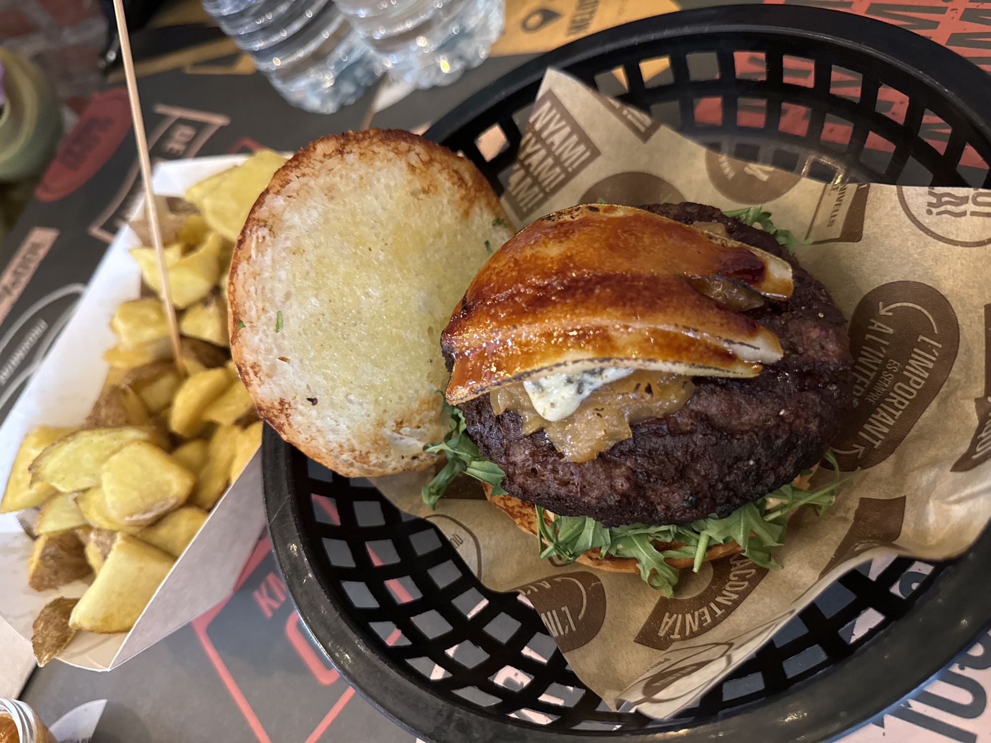 The image shows an open-faced burger in a black plastic basket, presented on brown paper. The burger consists of a patty, onions, blue cheese, caramelized pears, and arugula. Beside the burger is the top bun and a serving of potato wedges in a white paper container, impaled by a wooden skewer. Two water bottles are in the background. The brown paper in the basket features text in Catalan, indicating a Barcelona, Spain location, specifically naming the burger chain "KÖKÖ NIVELLS".