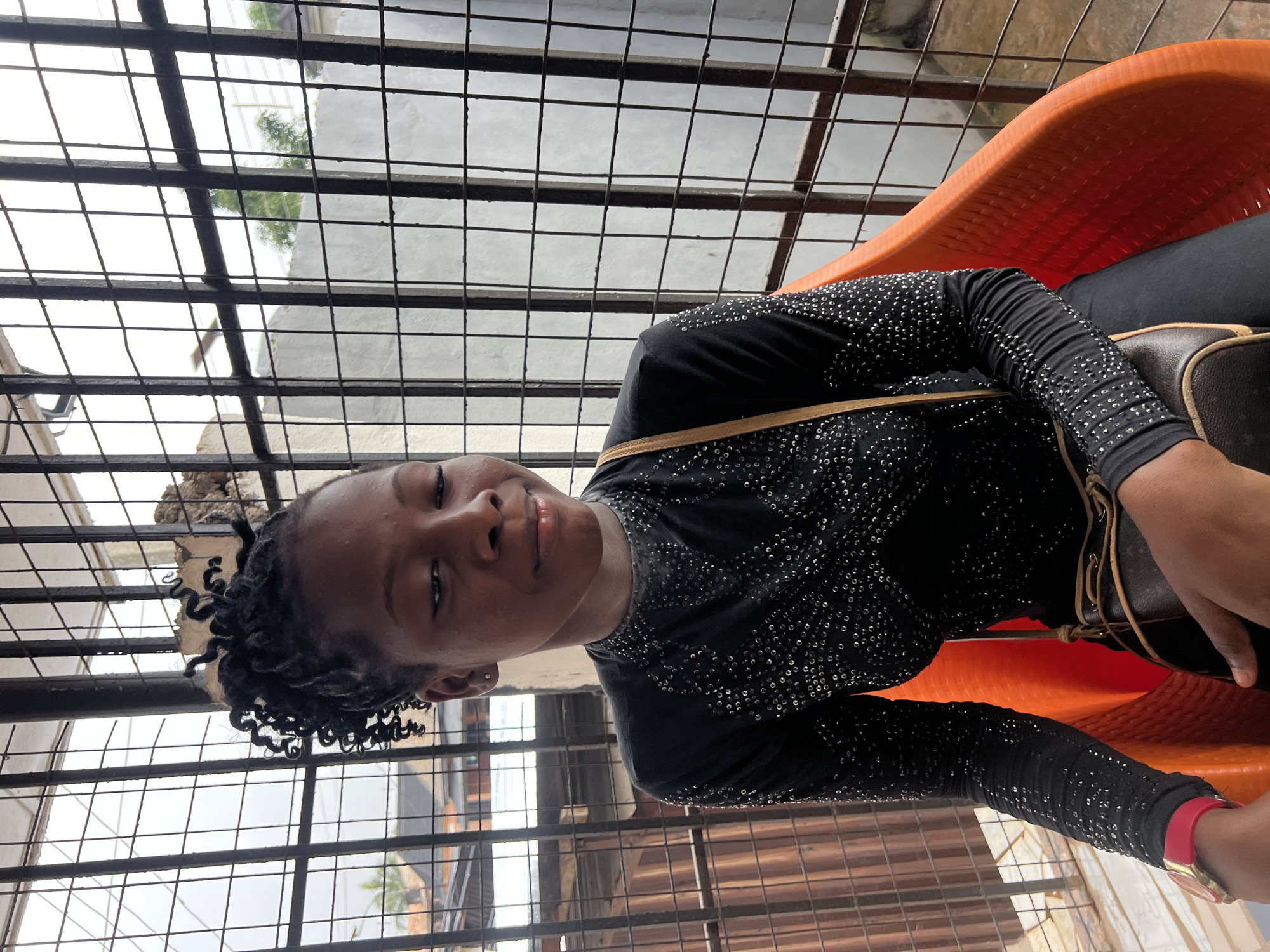 A young Black female is seated in a vibrant orange plastic chair, looking to her right with a slight smile. She has dark skin, black curly hair styled in a bun, and is wearing a black, long-sleeved top adorned with numerous small, reflective embellishments. A brown crossbody bag is slung over her shoulder, resting on her chest and lap. On her left wrist, she wears a watch with a red band and a gold-toned face. The background consists of a metal grate or fence, behind which is a building with corrugated metal siding on the left and a white wall with some greenery visible in the distance. The overall setting appears to be an outdoor or semi-outdoor area in Nkpor, Nigeria. The lighting suggests it is daytime, possibly overcast.