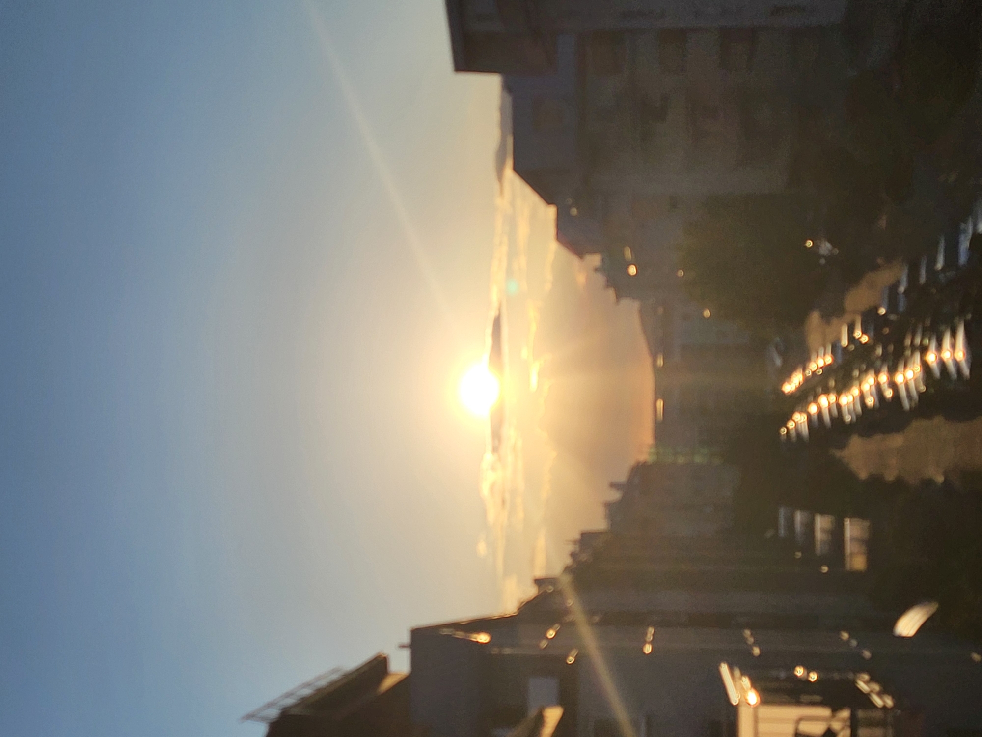This is an elevated, out-of-focus photograph of a city scene during either sunrise or sunset, with the location being Unknown, Unknown. The sky occupies the upper portion, exhibiting a gradient from light blue at the zenith to a vibrant yellow-orange closer to the horizon. A circular, intensely bright sun is centrally positioned on the horizon, emitting visible light rays and partially obscured by a horizontal formation of clouds that are illuminated by the sun's light.
Dark, blurred silhouettes of multi-story buildings flank a central street that recedes into the distance. On the left side, multiple structures with discernible rooftops and some bright, circular or elongated bokeh effects, indicative of light reflections or sources, are visible. The right side features similar indistinct building silhouettes.
The street below is lined with numerous parked vehicles, which appear dark and indistinct except for their upper surfaces (roofs and hoods). These surfaces intensely reflect the direct sunlight, creating a distinct, elongated pattern of bright, specular bokeh highlights that stretch towards the vanishing point of the street. The immediate foreground, including the street pavement and lower sections of the buildings, is deeply shadowed, appearing as dark masses against the brighter sky and car reflections. No human figures or specific identifying landmarks are discernible within the frame.