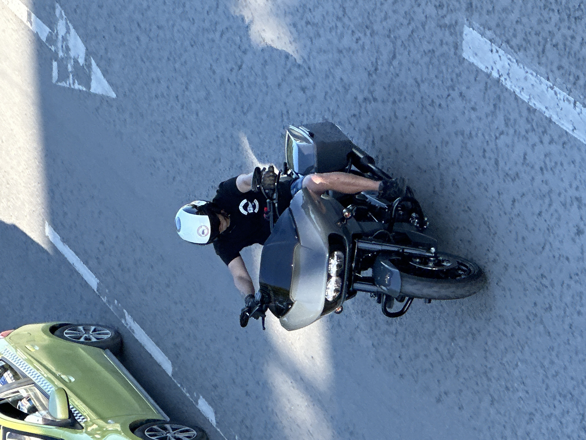 An overhead shot captures a man riding a motorcycle on an asphalt road. The man wears a white helmet and a black t-shirt with a circular logo. He is sitting on a dark-colored motorcycle. The road has white directional arrows and solid white lines. A green taxi cab is visible on the lower left. The location is Veliko Tarnovo, Bulgaria.