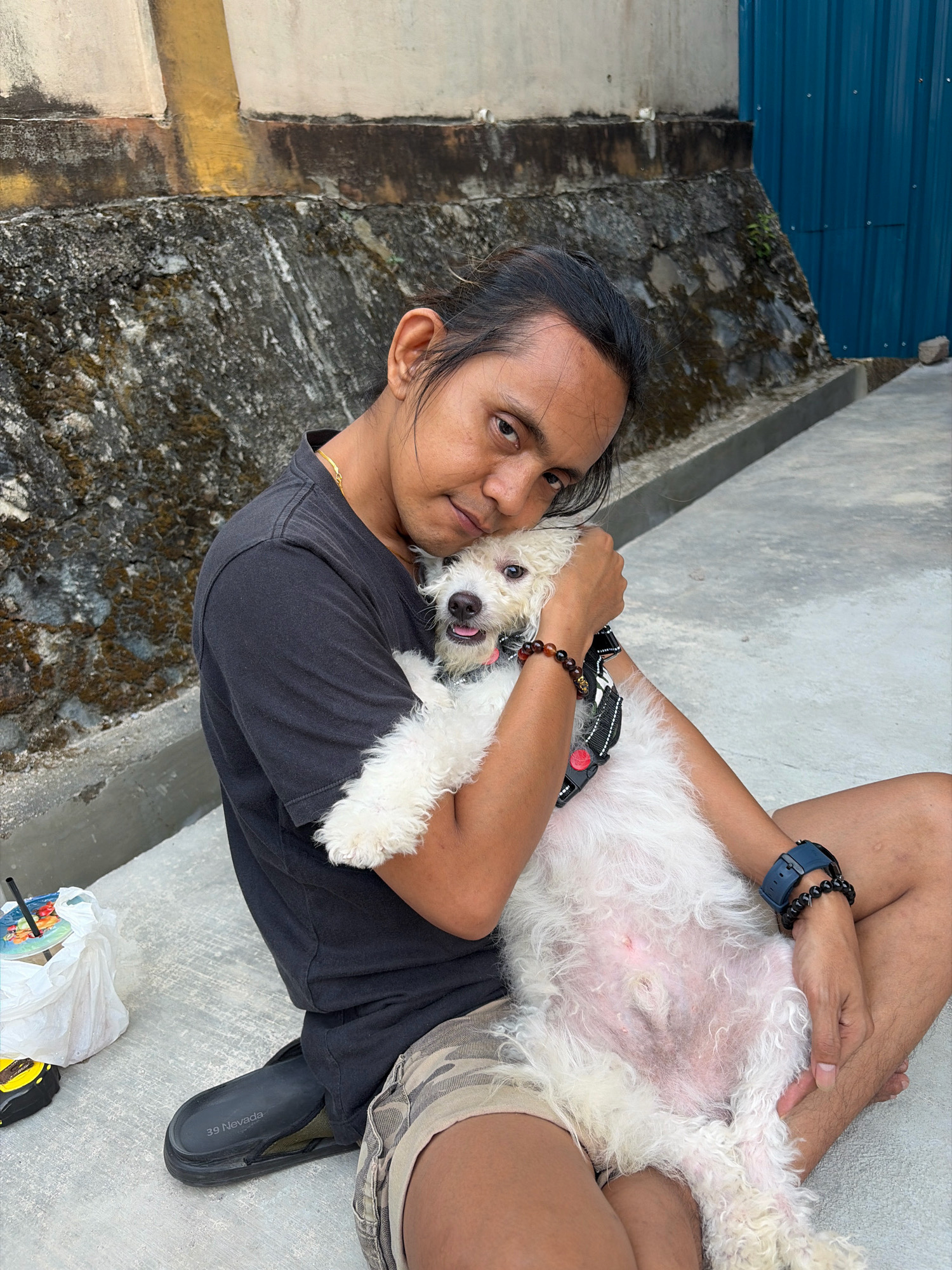A person is sitting on concrete, embracing a white poodle dog. The person, with dark hair pulled back, wears a black t-shirt and camouflage shorts. They have a gold chain around their neck and are wearing a dark blue watch and a beaded bracelet on their left wrist. The dog is nestled in the person's arms, with its mouth open and tongue slightly out. The background features a textured wall, a blue corrugated metal fence, and more concrete. To the left of the person, a white plastic bag with a drink and a yellow measuring tape are visible on the ground. The scene appears to be outdoors in an urban environment, possibly a courtyard or alleyway, in Tanjung Pinang, Indonesia.