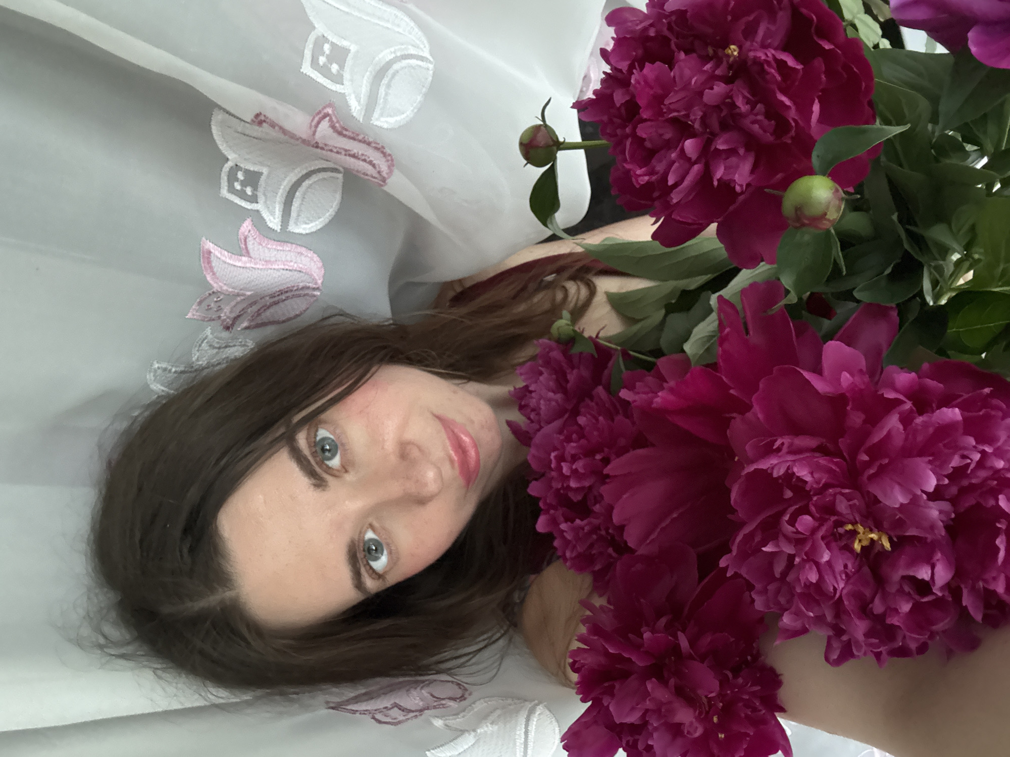 A woman with brown hair and blue eyes is holding a bouquet of deep pink peonies. She is wearing a dark red top. The background consists of a white sheer curtain with a floral pattern. The image appears to be taken indoors. There are no discernible city or environmental details.