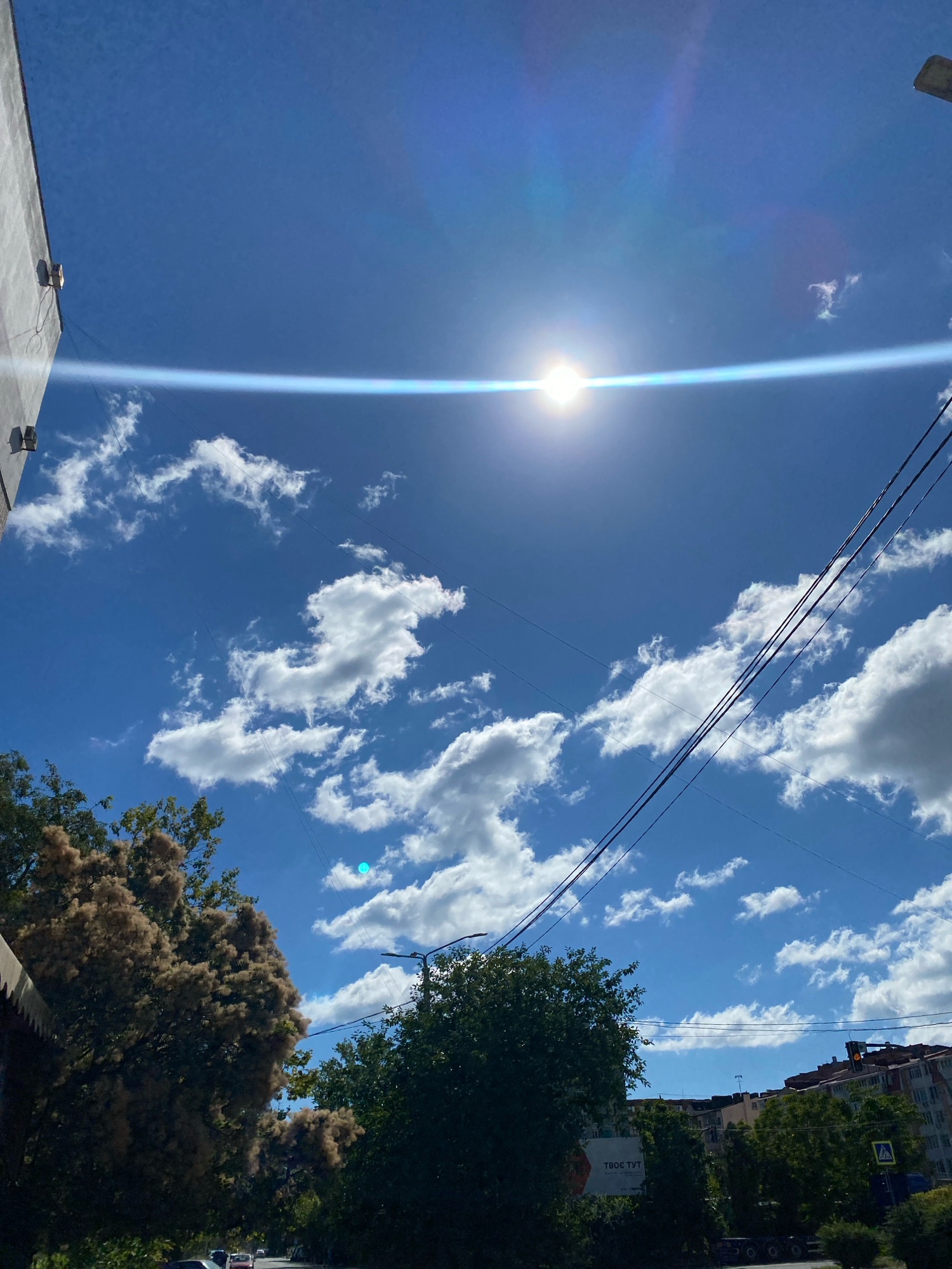 The image depicts a sunny day in Chornomorsk, Ukraine, characterized by a bright blue sky with scattered white cumulus clouds. The sun is visible near the top center of the frame, creating a lens flare that streaks across the sky horizontally. Overhead power lines traverse the sky diagonally from the upper right to the mid-left.
In the foreground, on the left, a building with a light-colored facade is partially visible. Two outdoor lighting fixtures are attached to this building. Below the building, trees with feathery, light brown foliage are prominent. To the right of these trees, another cluster of dark green trees and vegetation is visible, with a few buildings with red-tiled roofs peeking through. A street is visible at the bottom, with a few vehicles parked or driving. A traffic light displaying an orange signal is visible in the distance, above the buildings. A pedestrian crossing sign is also visible on the right side of the street.
