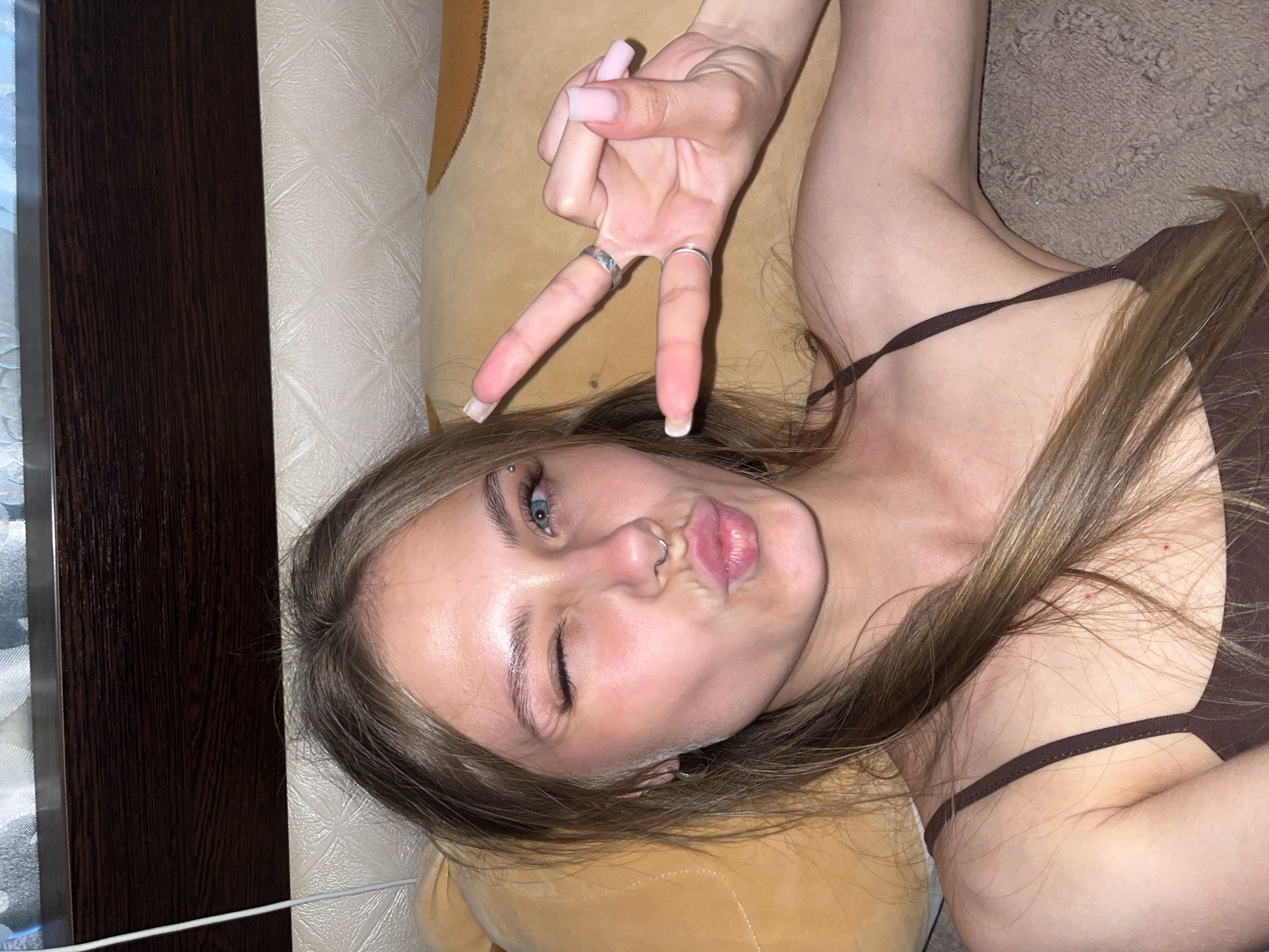 The image shows a young woman lying on her side, her head turned towards the camera. She is winking with her left eye and puckering her lips, giving a playful expression. Her right eye is open, revealing blue irises. She has light brown hair that falls around her face and shoulders. She is wearing a brown tank top. A silver ring is visible on her left index finger, and another on her right middle finger. Her fingernails are long and painted a pale pink. A nose ring and eyebrow piercing are visible. Her hand is held up in a peace sign. The background appears to be indoors, with a textured wall and a dark wooden surface on the left side. The lighting suggests it could be daytime or artificial light.