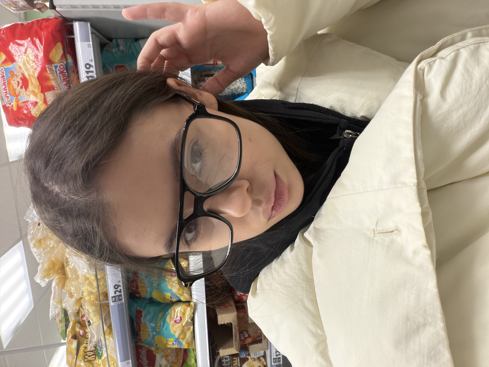 The image, oriented on its side, features a young person with long, dark brown hair and black-framed glasses, looking directly towards the camera with a neutral expression. One hand is raised, gently touching the hair above their ear. They are wearing a cream or off-white puffy jacket over a black inner garment.
The setting appears to be a grocery store or supermarket, with shelves of packaged food products visible in the background on both sides of the person. The products mostly consist of snacks like chips and crackers, indicating an aisle dedicated to such items. Bright overhead fluorescent lighting is reflected in the background.
Visible text on the product packaging, written in Cyrillic, includes "Солнышко" (Solnyshko, meaning "Sunshine") on a red bag, "ПОПКОРН" (Popcorn), "MIA BAR", and "МЮСЛИ" (Muesli, as identified from "ESLI"). Price tags are also visible, showing "119" and "29", likely indicating prices in rubles. The shelves are well-stocked, suggesting an active retail environment.