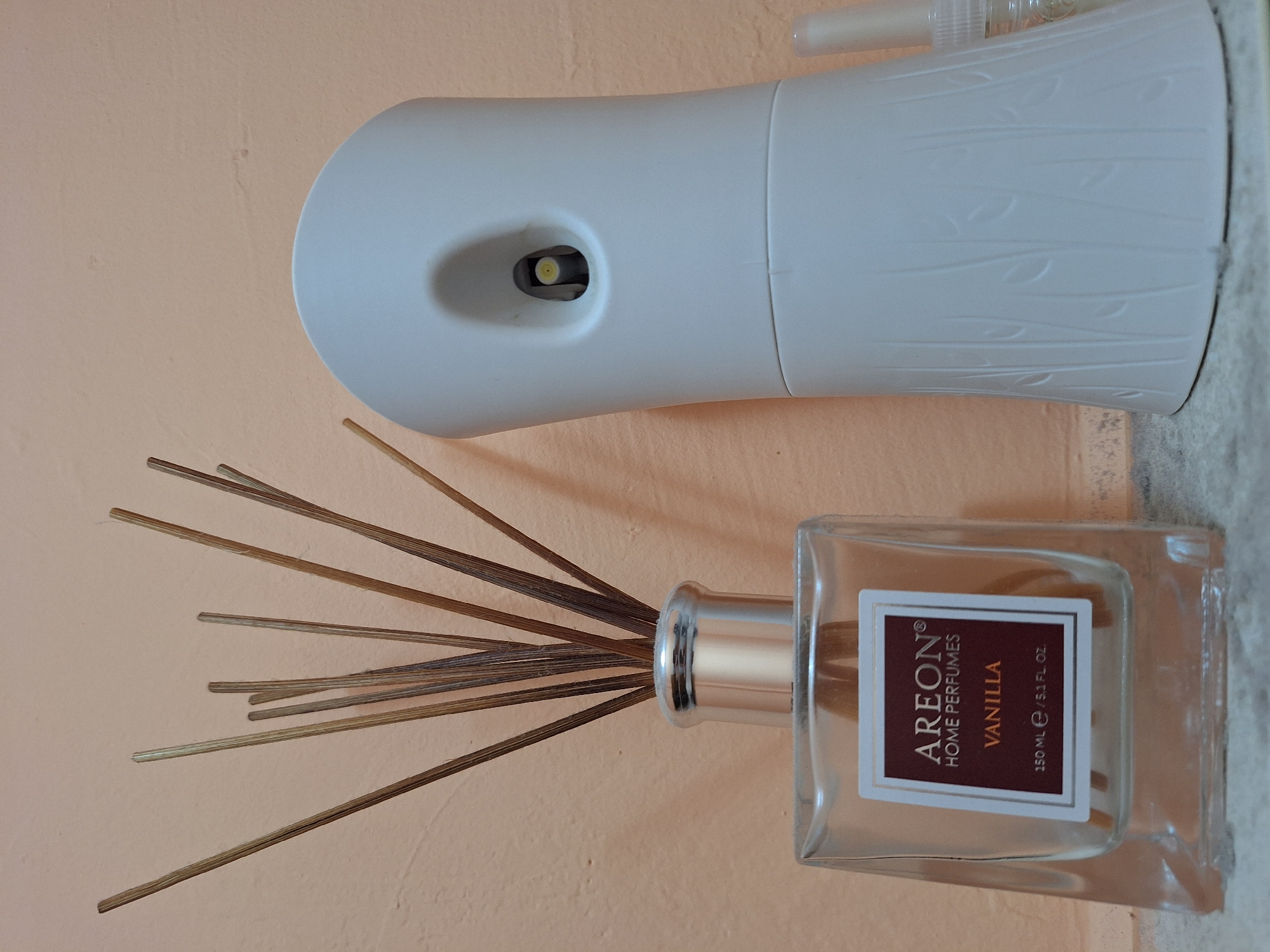 The image depicts a still life of home fragrance products against a peach-colored wall. On the left, a clear glass bottle with a silver cap contains several reed diffusers. The bottle has a label that reads "AREON HOME PERFUMES VANILLA 150 ML / 5.1 FL. OZ." To the right of the diffuser bottle is a white automatic air freshener with a textured design of vertical lines resembling reeds or leaves. A small spray bottle is partially visible to the far right of the image. The foreground shows a speckled gray surface. The scene is indoors, with no indication of a specific location within Varna, Bulgaria.