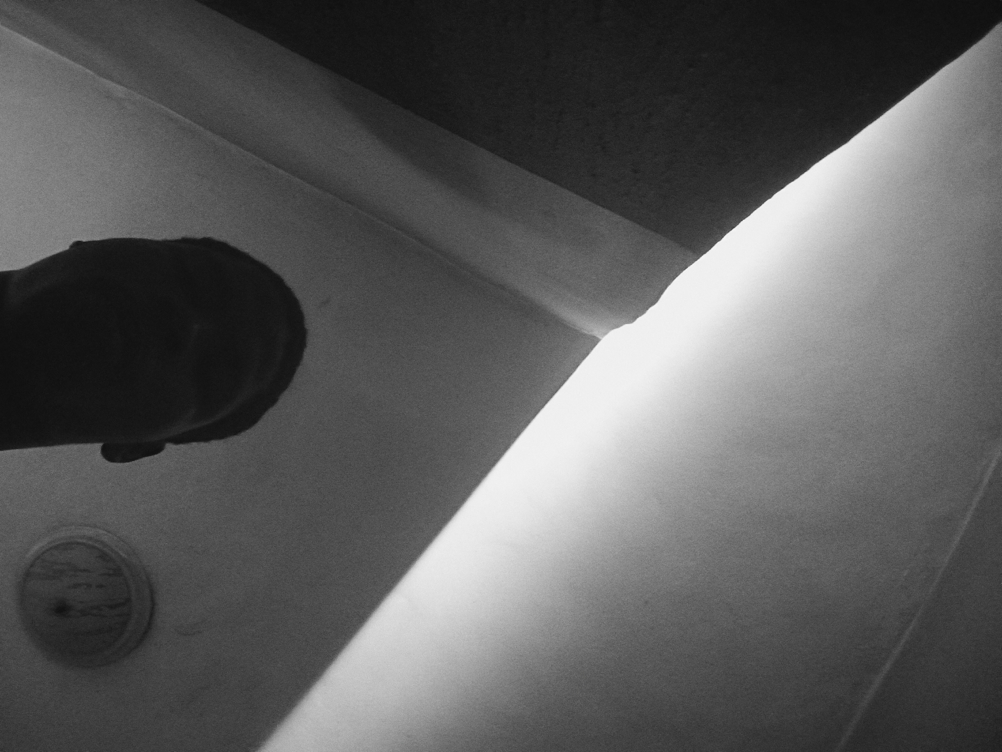 A high-contrast, black and white photograph captures an interior scene, dominated by strong directional light and shadow. On the left side of the frame, a dark, silhouetted form, resembling a human arm or the back of a head/shoulder, extends from the upper left, partially obscured. Below this form, towards the bottom left, is a round, textured object, possibly made of wood or metal, with a central detail that could be a screw or knob.
The remainder of the image is defined by architectural elements. A prominent, intensely bright diagonal shaft of light cuts across the frame from the upper right corner to the lower left, creating a stark division between illuminated surfaces and deep shadows. This light emphasizes the texture of what appear to be walls or structural beams, which meet at sharp angles in the upper and middle sections of the image. The surfaces are generally smooth but show a noticeable graininess typical of film photography or high-ISO digital images.
No specific activity is taking place, and the scene is still and contemplative. The strong light suggests daylight entering from an unseen source, such as a window or doorway, implying it could be morning or afternoon. There are no overt location cues beyond the architectural style, which is simple and functional. No text is visible within the image. The overall impression is one of minimalist forms, strong composition, and a focus on the interplay of light and shadow within an anonymous indoor space in Nkpor, Nigeria.