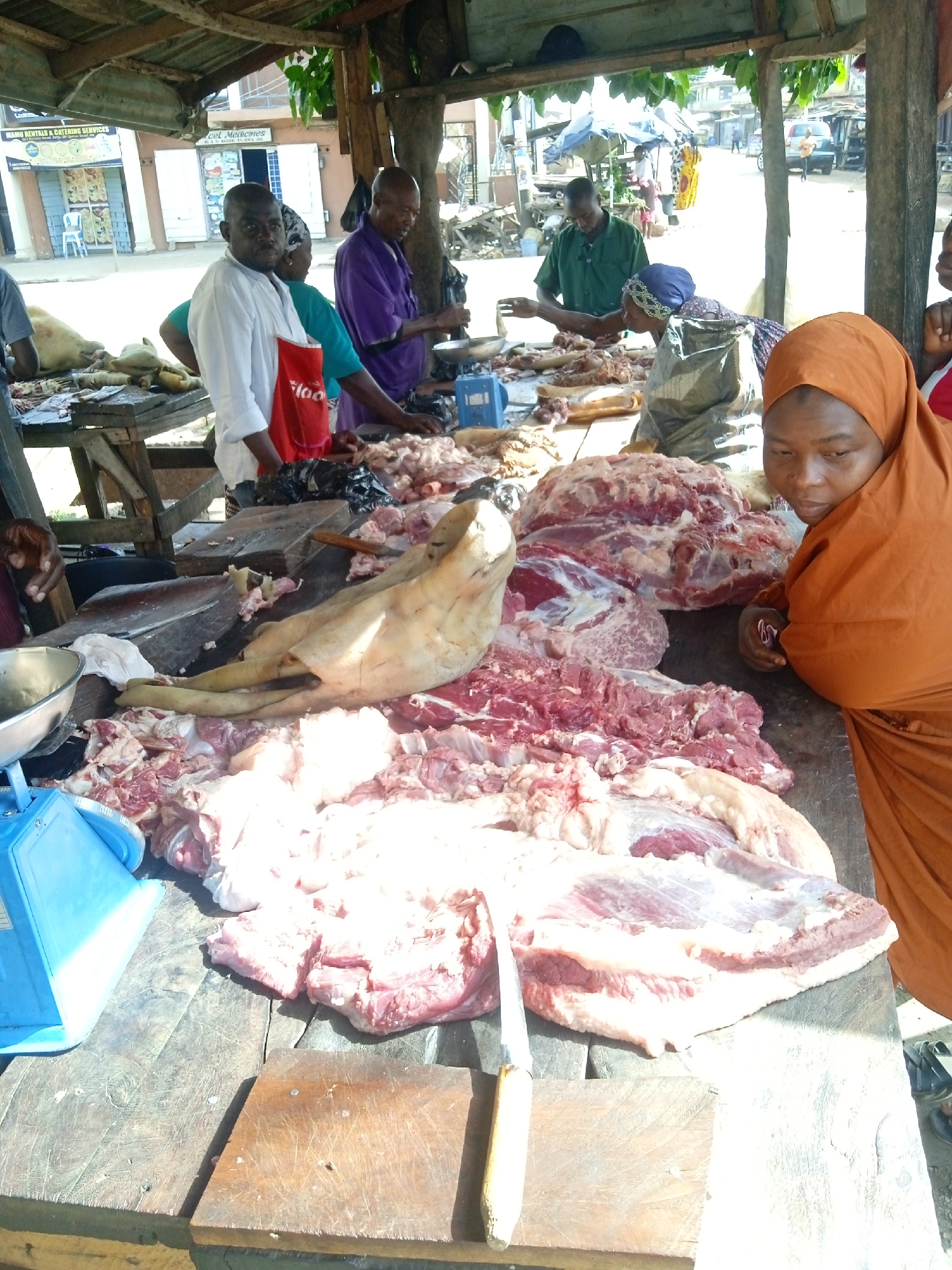The image depicts a bustling outdoor meat market in Jos, Nigeria. Several butchers are seen working behind a large wooden table laden with various cuts of raw meat, including what appears to be a pig's head. One woman wearing an orange hijab is standing near the table looking at the camera, possibly a customer. A blue weighing scale is visible on the table. Behind the table are several men, some of whom are cutting and handling the meat. In the background, there are buildings, street scenes, and people moving about. The market is covered by a corrugated metal roof.