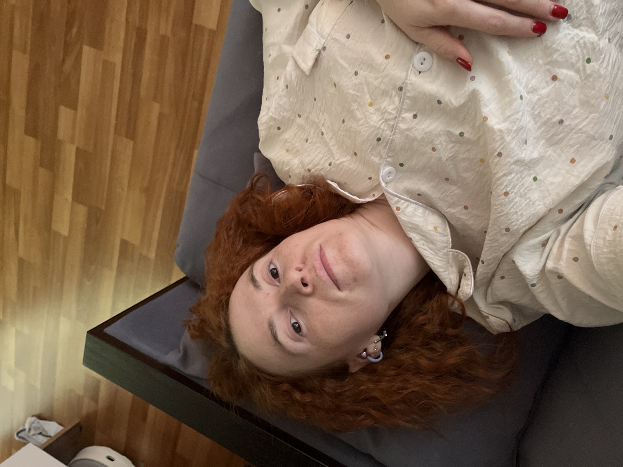 A woman with red hair, wearing a polka dot pajama top with red nail polish, is reclining on a grey upholstered platform. The platform is positioned near a wooden floor. The location is unknown and the presence of the city of Chaikovsky, Russia cannot be determined from the image.