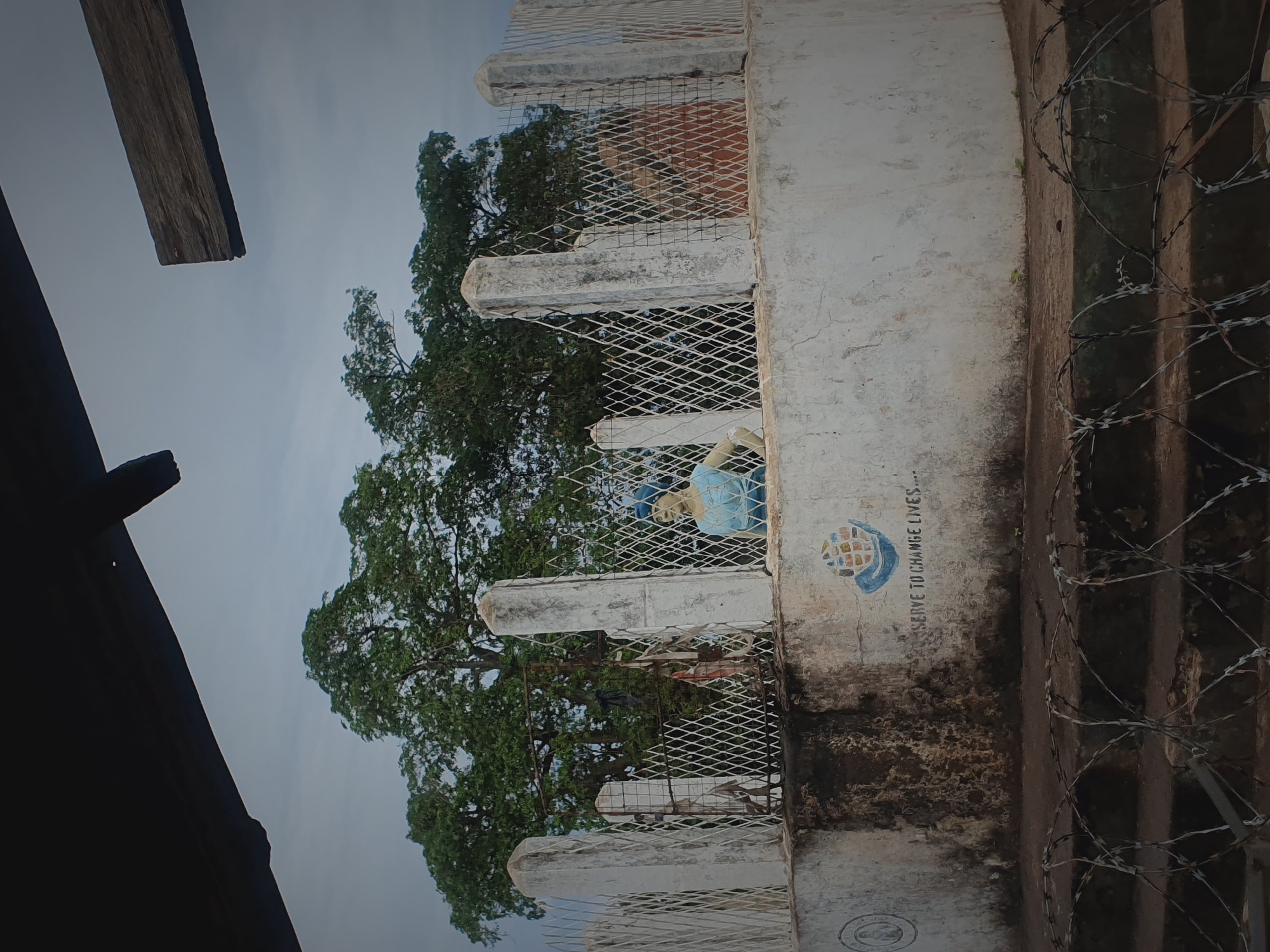 The scene is an outdoor setting in Idah, Nigeria, depicted under an overcast sky. The upper portion of the image shows a dark, solid architectural beam on the left and a wooden plank on the right, partially obscuring the sky.
In the midground, a weathered, circular concrete wall or base, painted white and showing significant dark staining and dirt, is prominent. This wall supports a white metal fence composed of diamond-patterned mesh panels fixed to multiple concrete posts. Behind the fence, slightly left of center, the light-blue colored bust of a human figure, wearing a head covering and a simple garment, is visible. Near the center of the fence, a seemingly damaged gate with some dark fabric-like items hanging on it is present. On the exterior surface of the concrete wall, to the right, a painted emblem depicts two hands cupping a multi-colored globe, with the partial inscription "...SERVE TO CHANGE LIVES..." below it.
Behind the concrete structure and fence, a dense canopy of large green trees forms the background. The immediate foreground is dominated by multiple coils of razor wire, spread across a set of low, dark concrete steps or a raised platform.