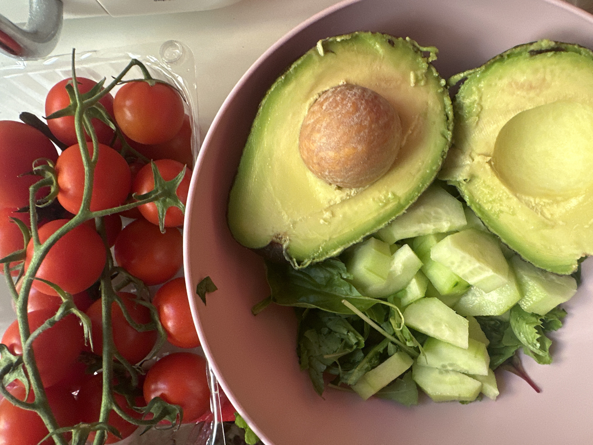 This is a top-down, close-up media file showing a collection of fresh ingredients for a salad. The main subjects are cherry tomatoes in a clear plastic container and a salad bowl containing a halved avocado, chopped cucumber, and leafy greens. The tomatoes are a vibrant red and are still on the vine. The avocado is ripe with a large pit in one half. The cucumber is cut into bite-sized pieces, and the greens appear to be a mix of lettuce and possibly other herbs.
The setting is a bright, indoor environment, likely a kitchen counter, given the presence of what appears to be a faucet or sink fixture in the upper left corner. The lighting suggests it is daytime. There is no visible text in the media file. The overall impression is one of freshness and healthy eating, possibly preparing a meal or snack. The location context of Milan, Italy, is not visually evident from the image itself.