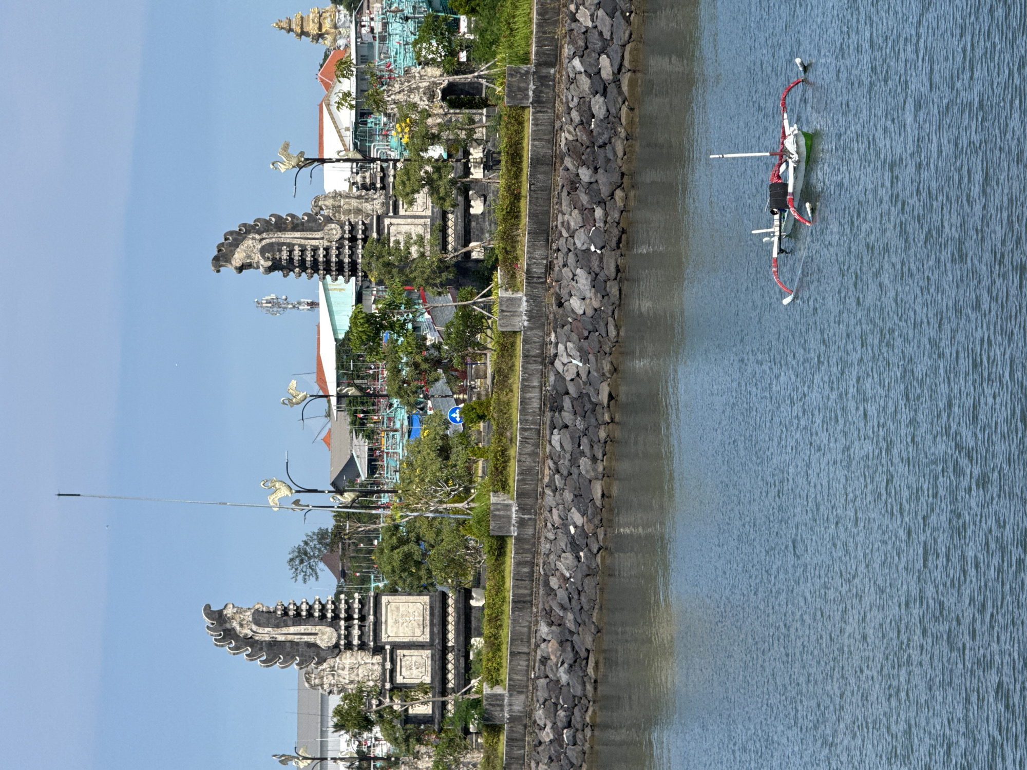 The image depicts a coastal scene in Denpasar, Indonesia, featuring traditional Balinese architecture and a small outrigger boat in the water. A stone seawall or embankment separates the land from the water. The architecture includes ornate gates and decorative elements, suggesting a temple or public space. Lush greenery is present along the shoreline. The boat is decorated with red elements. The sky is clear.