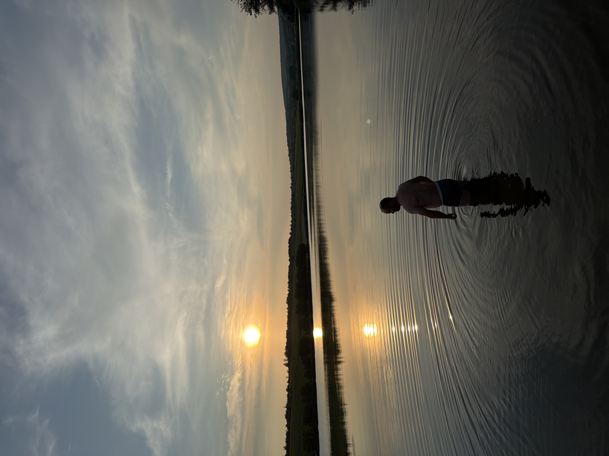 A shirtless man stands in shallow water facing away from the camera, with his reflection visible in the rippling surface of a lake. The sun, low in the sky, casts a warm glow and reflects on the water. The background shows a tree-lined shore and rolling hills. The sky is filled with wispy clouds.