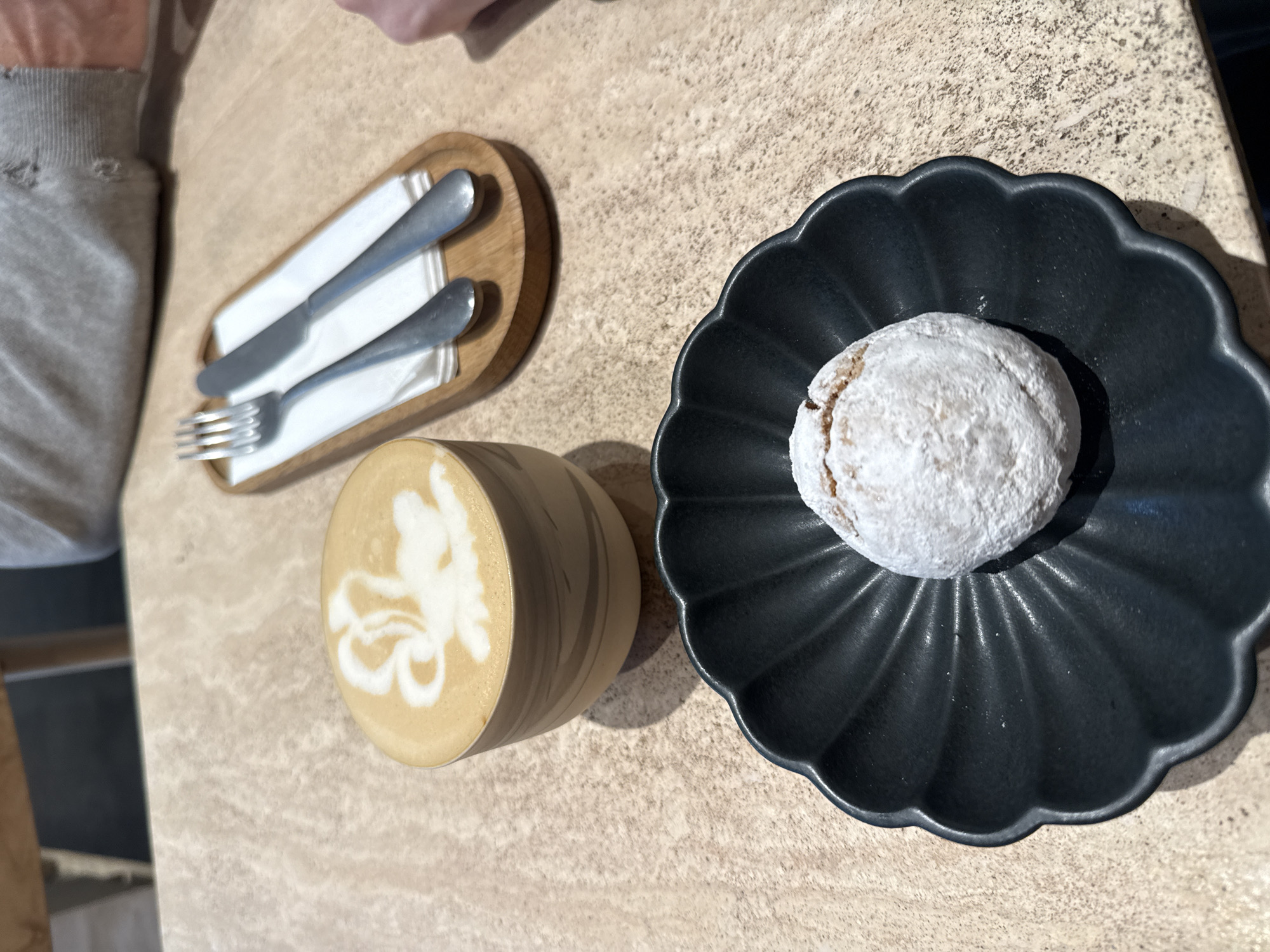 The image shows a coffee drink with latte art in a cylindrical cup, a cookie covered in powdered sugar on a black decorative plate, and silverware on a small wooden tray. The objects are arranged on a tan-colored surface. The location is unknown and there is no indication it's Moscow, Russia.