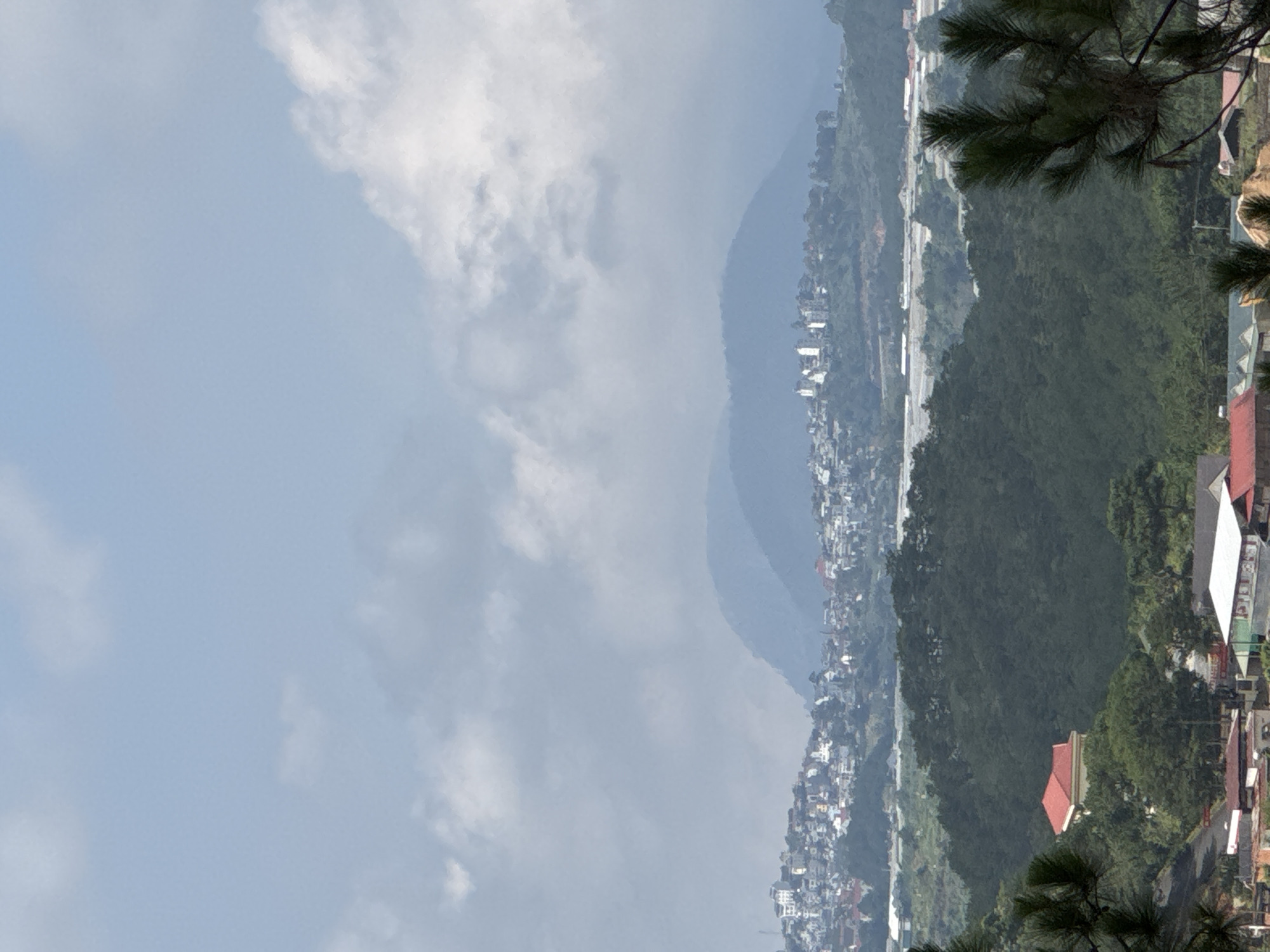 The image depicts a hillside town or city. Buildings are densely packed along the slopes of mountains, interspersed with green vegetation. A road runs through the built-up area, and in the foreground, pine trees frame the view. The sky is overcast, suggesting a cloudy or possibly hazy day. Due to the lack of unique identifiers in the image, it cannot be confirmed that the scene is located within Lạc Dương, Vietnam.
