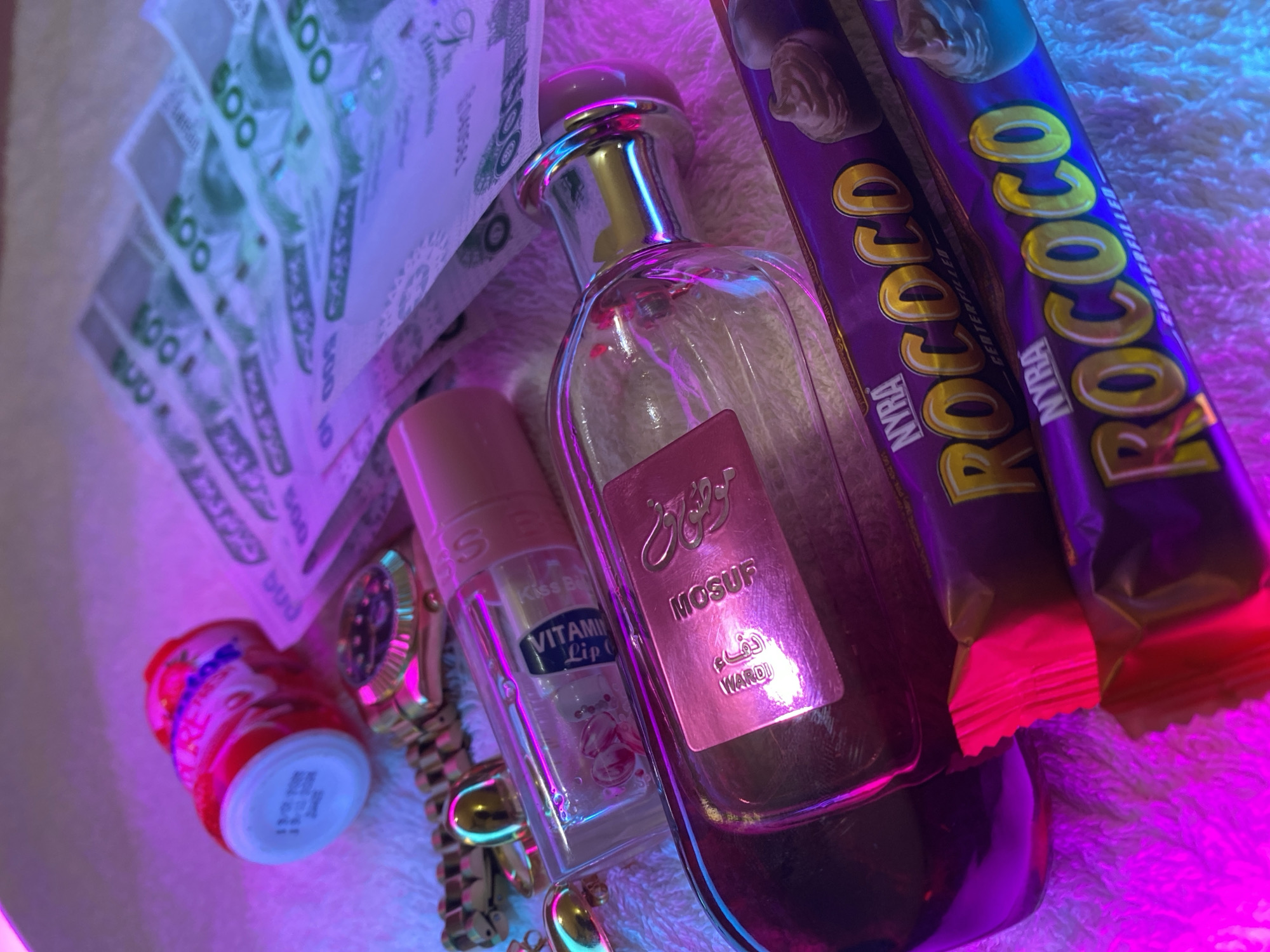 A vibrant flat lay composition showcases an array of personal items and currency, set against a light-colored, textured surface under strong pink and purple lighting in Maigizo, Nigeria.
In the upper left, several Nigerian 500 Naira banknotes are fanned out, prominently displaying the number "500." Central to the image is a clear glass perfume bottle with a golden cap and a pink label that reads "MOSUF" and "WARDI," along with Arabic script. To its left, a smaller clear bottle with a pink cap is identified as "Kiss Bi VITAMIN Lip Oil." A gold-toned wristwatch with a dark face and metallic links is partially visible beneath these items.
On the right side of the frame, two purple-wrapped chocolate bars are stacked, both clearly labeled "ROCOCO" in yellow lettering, with additional text indicating "NYRA" and "CENTERFILLED." In the lower-left corner, a small, red and white cylindrical container is visible, though most of its text is obscured. The artificial, neon-like lighting casts a strong purple and pink glow over all objects, contributing to a stylish and vivid aesthetic.