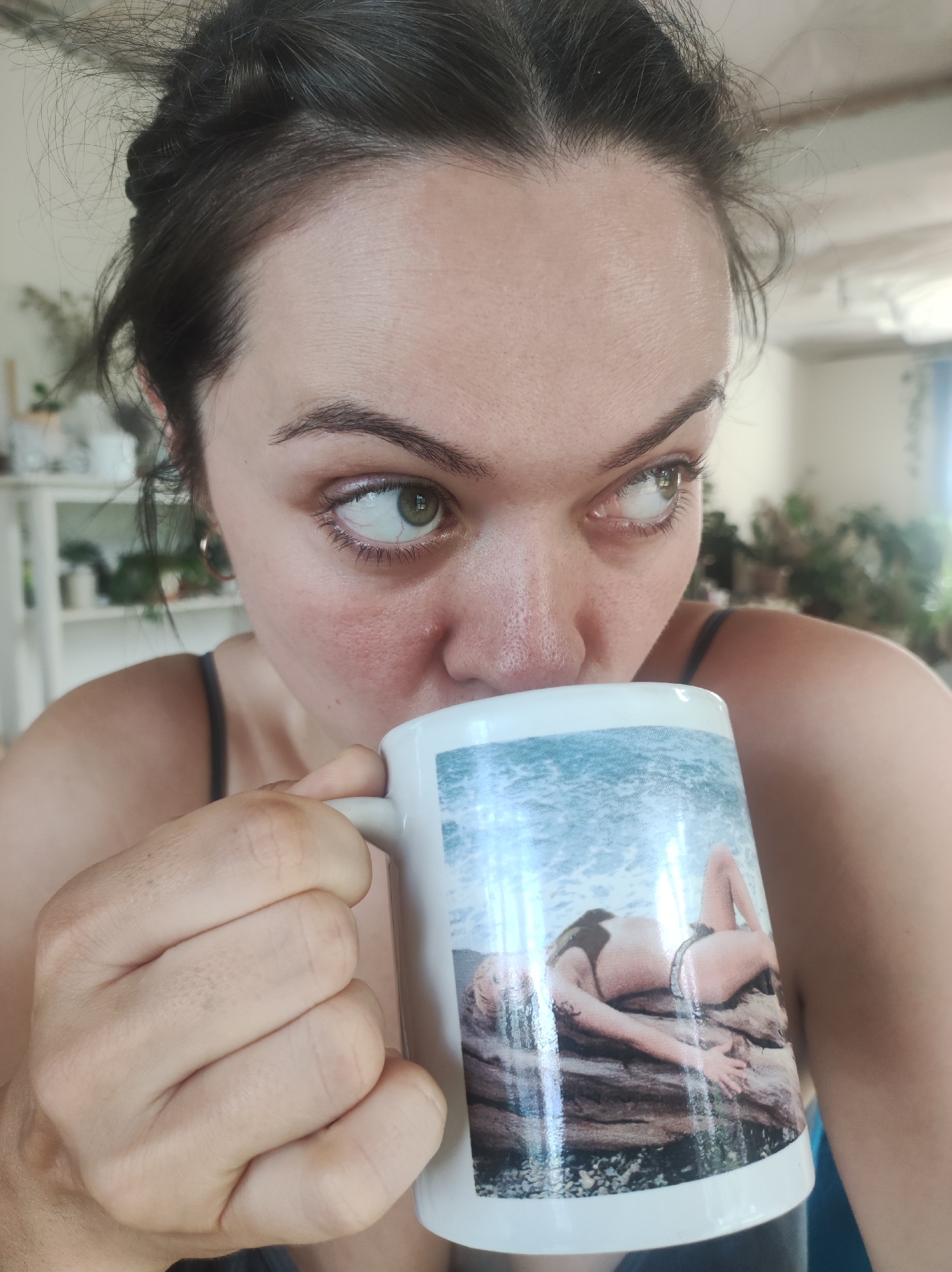 A close-up, indoor photograph taken in the city of Фоки, Russia, depicts an adult female drinking from a white ceramic mug. The female, positioned centrally, has dark hair loosely pulled back and green eyes looking towards the right side of the frame. Her right hand, with fingers curled, holds the mug to her mouth. The mug features a printed image of a light-haired individual in dark swimwear reclining on a piece of driftwood or rock at a waterfront. The background is softly out of focus, showing a residential interior with white shelving units displaying potted plants and other objects on the left, and indistinct household items and greenery on the right.