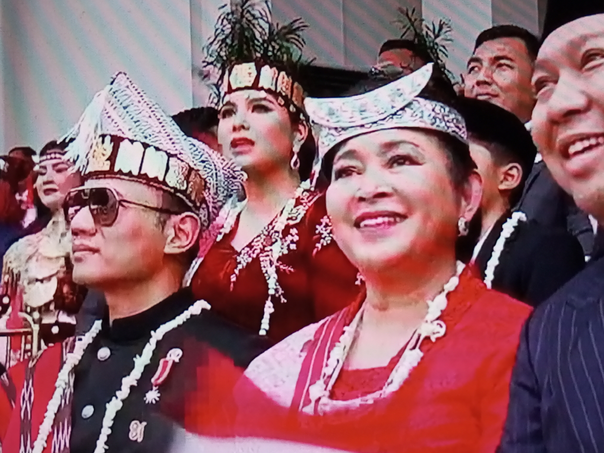 The image shows a close-up of several people at an event, likely in Banyudono, Indonesia, judging by the traditional attire and the presence of what appear to be Indonesian flags or banners. In the foreground, a man on the left wears a black jacket with white trim and a highly decorative, conical hat with gold embellishments. He is also wearing dark sunglasses and has a medal pinned to his jacket. To his right, a woman is smiling, her head adorned with a similar decorative hat with a feathered plume. She is wearing a red garment with intricate white embroidery. Behind them, other individuals are visible, including a woman in a red dress with a feathered headdress, and men in formal attire. The overall impression is of a formal or celebratory occasion, possibly a cultural ceremony or a public event.