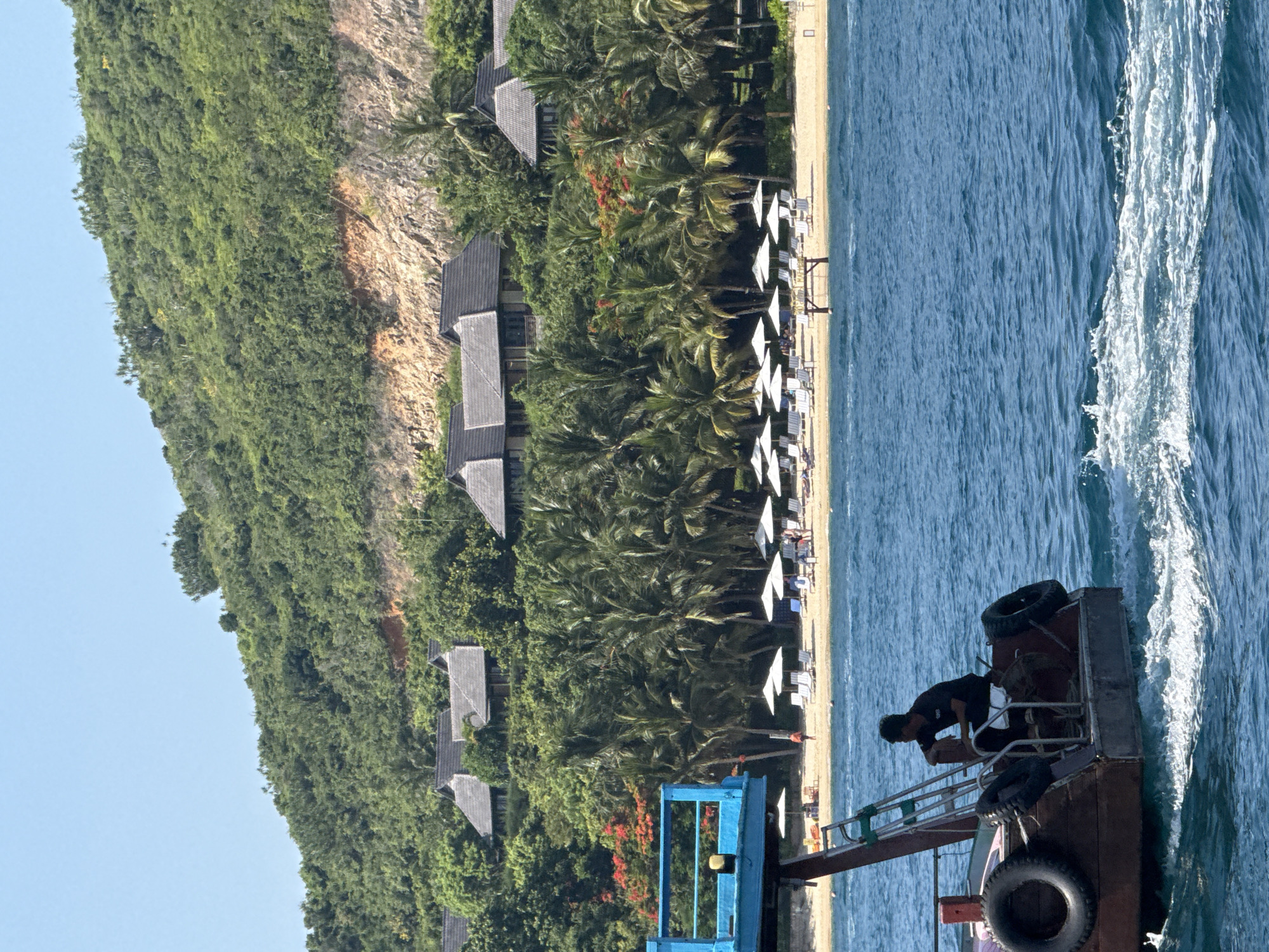 A boat with a single person visible on it travels on blue water, leaving a wake. In the background, a sandy beach lined with palm trees and white umbrellas stretches out. Behind the beach, a hillside covered in dense green foliage rises, dotted with a cluster of houses with dark, sloped roofs. The sky is clear and blue. The scene appears to be the coast of Nam Nha Trang Ward, Vietnam.
