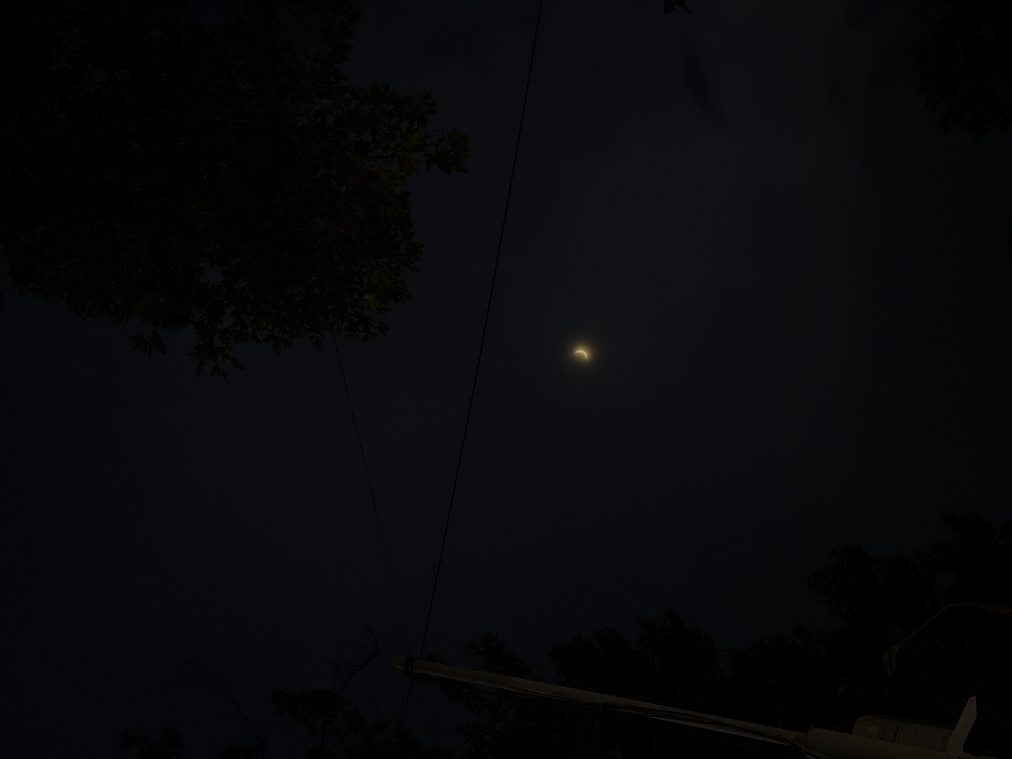 The image captures a crescent moon against a dark, cloudy sky, likely at dusk or night. Silhouetted against the sky are the dark shapes of trees and foliage in the foreground and background, suggesting an outdoor setting, possibly a park or a residential area with trees. A thin, dark power line runs diagonally across the image, adding a man-made element to the natural scene. In the lower right portion of the image, a faint, blurry object, possibly a part of a structure or equipment, is visible. The overall mood of the image is serene and slightly mysterious due to the low light and the prominent moon. No people are visible in the frame, and there is no visible text. The scene implies a quiet moment in nature, possibly in Chirchik, Uzbekistan, as suggested by the location context. The weather appears to be overcast, obscuring any stars and giving the sky a uniform dark tone. The bright crescent of the moon is the focal point, casting a subtle glow that illuminates a small portion of the dark sky.