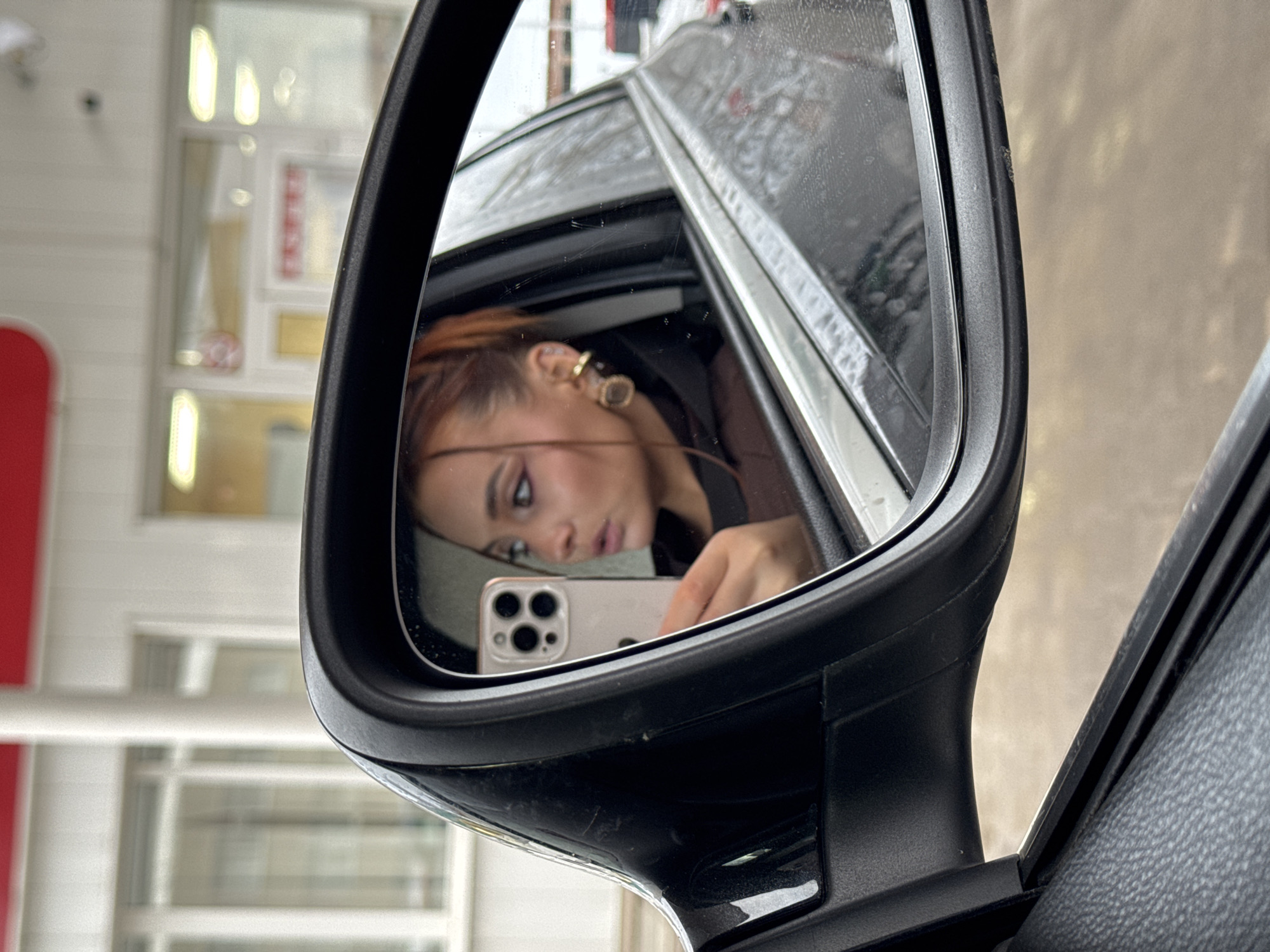 The image depicts a close-up view from inside a dark-colored vehicle, focused on the driver's side rearview mirror. Reflected in the mirror is a young adult female with reddish-brown hair, styled partially pulled back with a few strands framing her face. She has prominent dark eye makeup, including winged eyeliner, and multiple gold-toned earrings on her left ear, specifically a helix cuff and a larger circular dangle earring. Her lips are slightly parted, suggesting an expression of surprise or engagement. She is holding a light-colored smartphone, identifiable as an iPhone with a triple camera system, in her right hand, positioned as if taking a photograph or viewing the screen. She wears a dark garment, possibly a brown or black hoodie. The car's side window, visible in the reflection, shows streaks of dirt or moisture.
The background, visible both through the car window and reflected in the mirror, shows a commercial building. This structure features white walls with a distinct red fascia or trim along its upper section. Rectangular windows are visible. A blurred sign with Russian text "ПРОДУКТЫ" (PRODUKTY), meaning "GROCERIES" or "PRODUCTS," is discernible on the building, indicating a retail establishment, likely a grocery or convenience store. The overall lighting suggests an overcast environment. The scene is located in a commercial area within the city of Chaikovsky, Russia, adjacent to a store, possibly a convenience shop associated with a gas station, given the building's architecture and red-and-white color scheme.