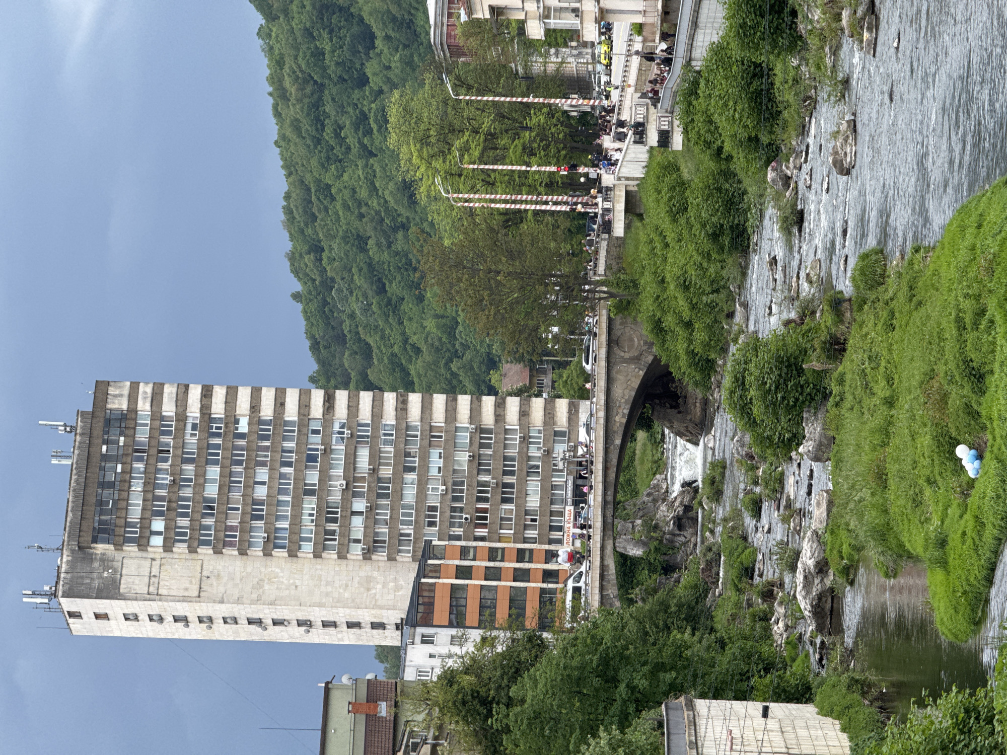 The image depicts a scene in Gabrovo, Bulgaria, featuring a multi-story building next to the Yantra River. The river flows over rocks and through greenery, with a bridge arching across it. The bridge has people crossing, and there are red and white striped barriers on the adjacent street. A forested hillside rises in the background. The sky is overcast. In the foreground, there are blue and white balloons. The building is a high-rise, constructed of concrete and windows.