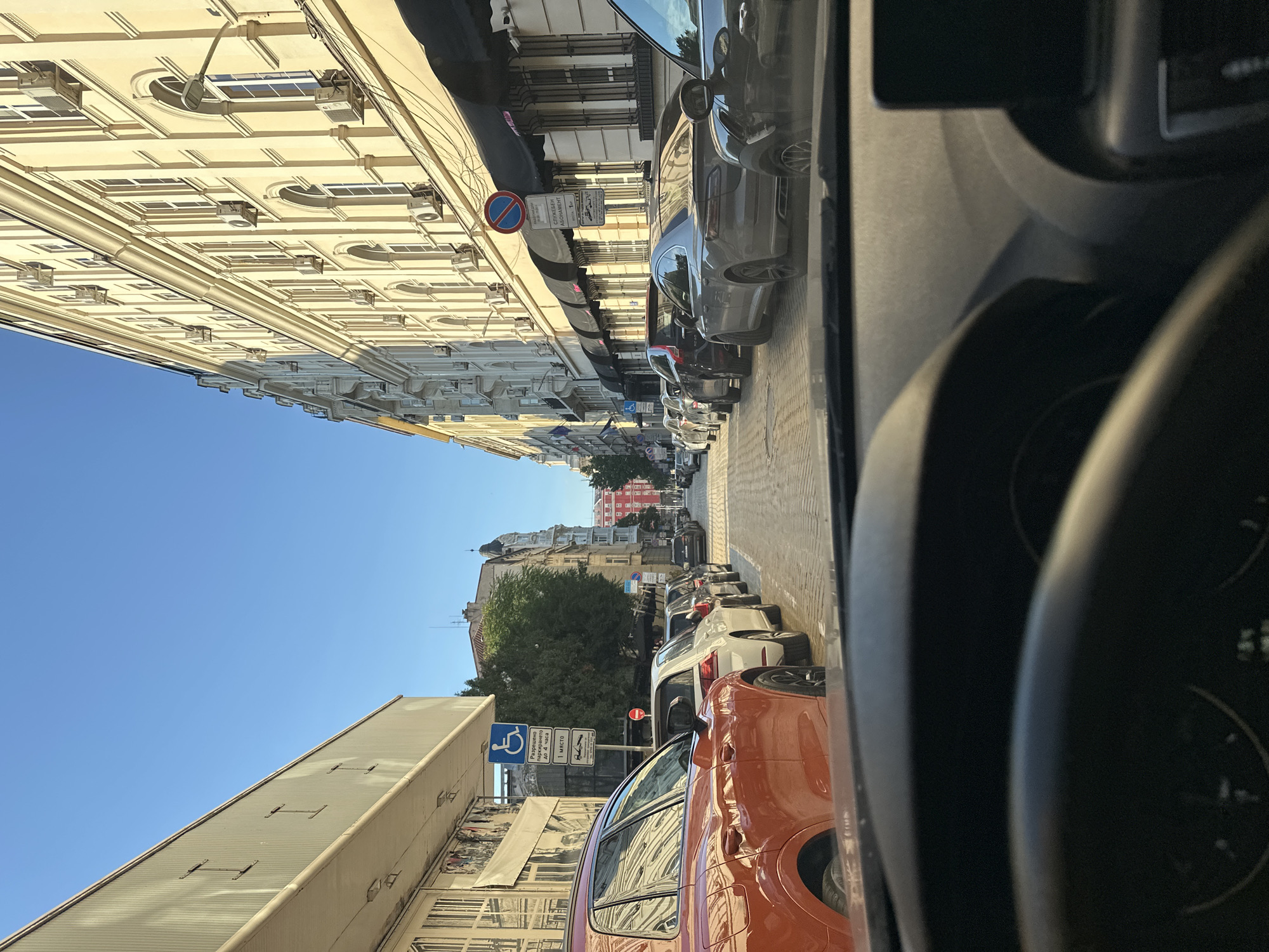 The image, taken from inside a vehicle, shows a street in Sofia, Bulgaria. The view is looking forward down a narrow street lined with buildings and parked cars. On the left side of the street, there is a light-colored building with large windows and some signage. A blue parking sign for disabled individuals is visible. On the right side of the street, a tall, ornate building with many windows and air conditioning units stands. Several cars are parked along both sides of the street. The street itself is paved with cobblestones. The sky is clear and blue, indicating daytime. The interior of the car, including a portion of the dashboard and steering wheel, is visible in the foreground.