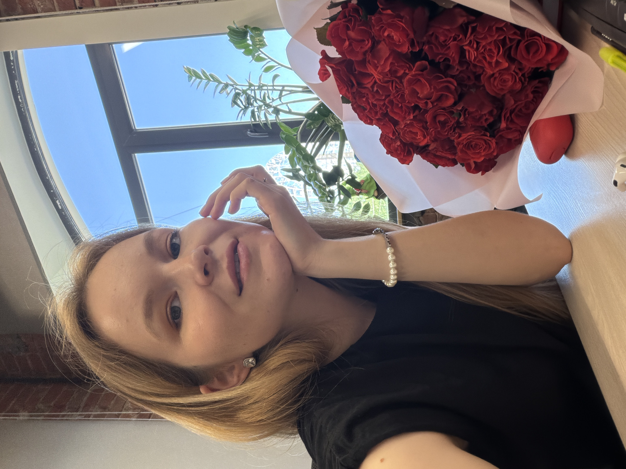 A young woman with fair skin and light blonde hair is posing for a selfie indoors. She is wearing a black shirt and a pearl bracelet on her left wrist. Her left hand is gently touching her cheek. She has a small, square earring in her left ear.
To the right of the woman, there is a bouquet of red roses wrapped in white paper on a light-colored surface, likely a desk or table. Behind the woman and the roses, a window reveals a clear blue sky and a green plant, suggesting it's daytime.
Although a specific location cannot be determined from this picture, the query specified the city of Izhevsk, Russia.
