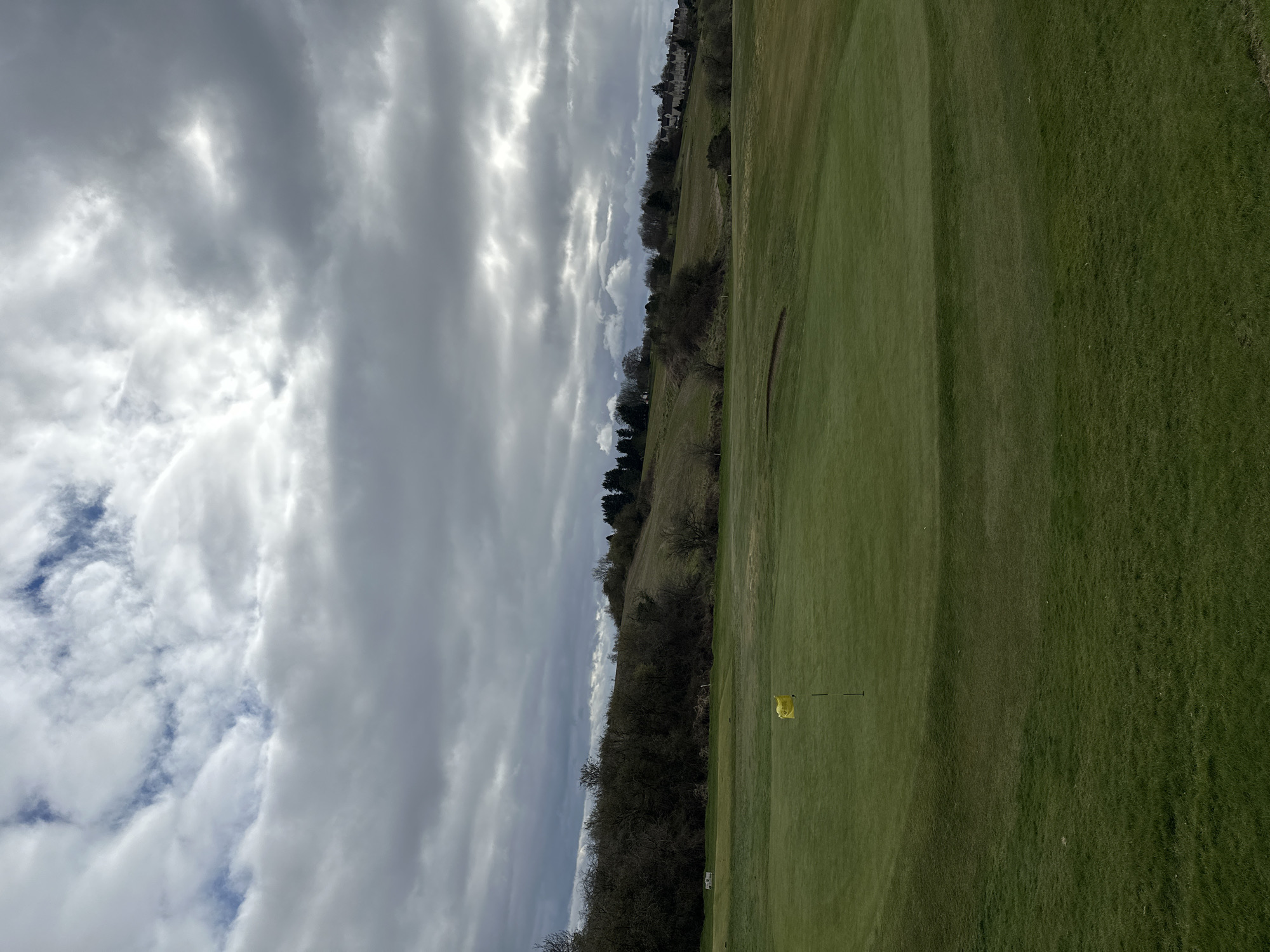 The image shows a golf course in Dunfermline, United Kingdom. The sky is overcast with heavy clouds. To the right is a green expanse of grass, indicating the fairway. In the middle of the fairway, a yellow flag marks a hole. A wooded area borders the left side of the fairway, transitioning into a slightly elevated area with sparse trees. In the distance, buildings are visible. The photo is taken on an overcast day.