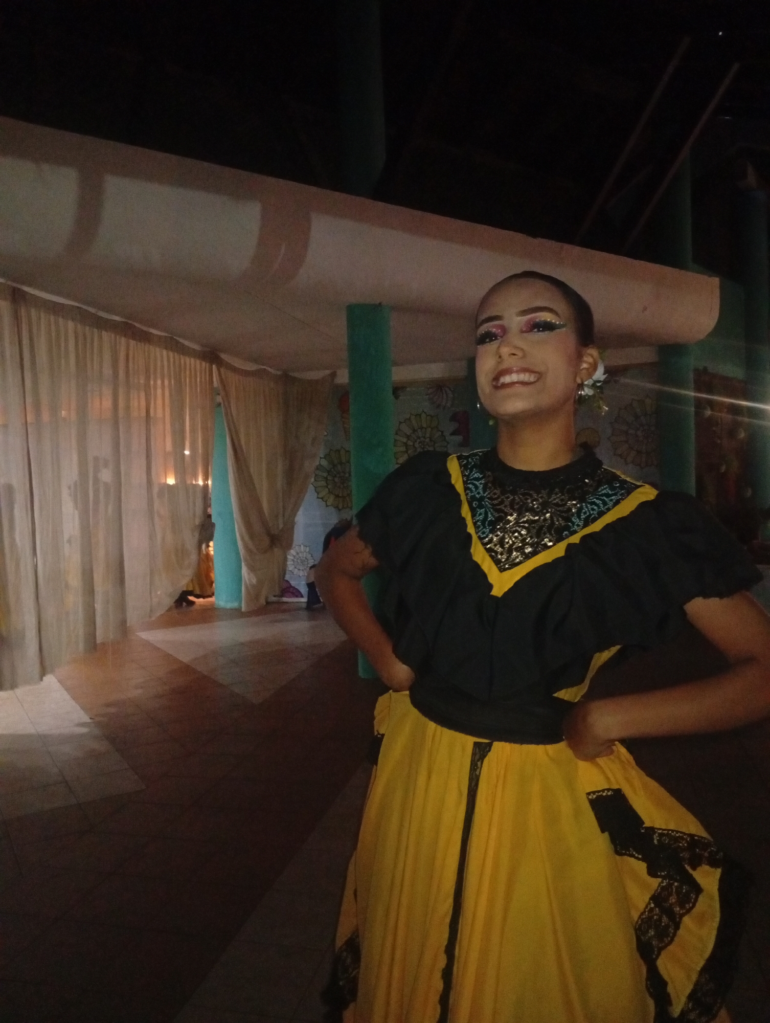 The image shows a young woman standing indoors. She is wearing a traditional dress, likely for a cultural performance. The dress has a black top with yellow trim and a yellow skirt with black lace accents. She has her hands on her hips and is smiling. Her makeup is vibrant, with colorful eyeshadow and glitter. She has a flower in her hair. The background appears to be a room or covered patio with curtains, pillars, and some decorative elements on the walls. The floor has a patterned tile design. This is likely in Playa El Agua, Venezuela, as specified in the prompt.