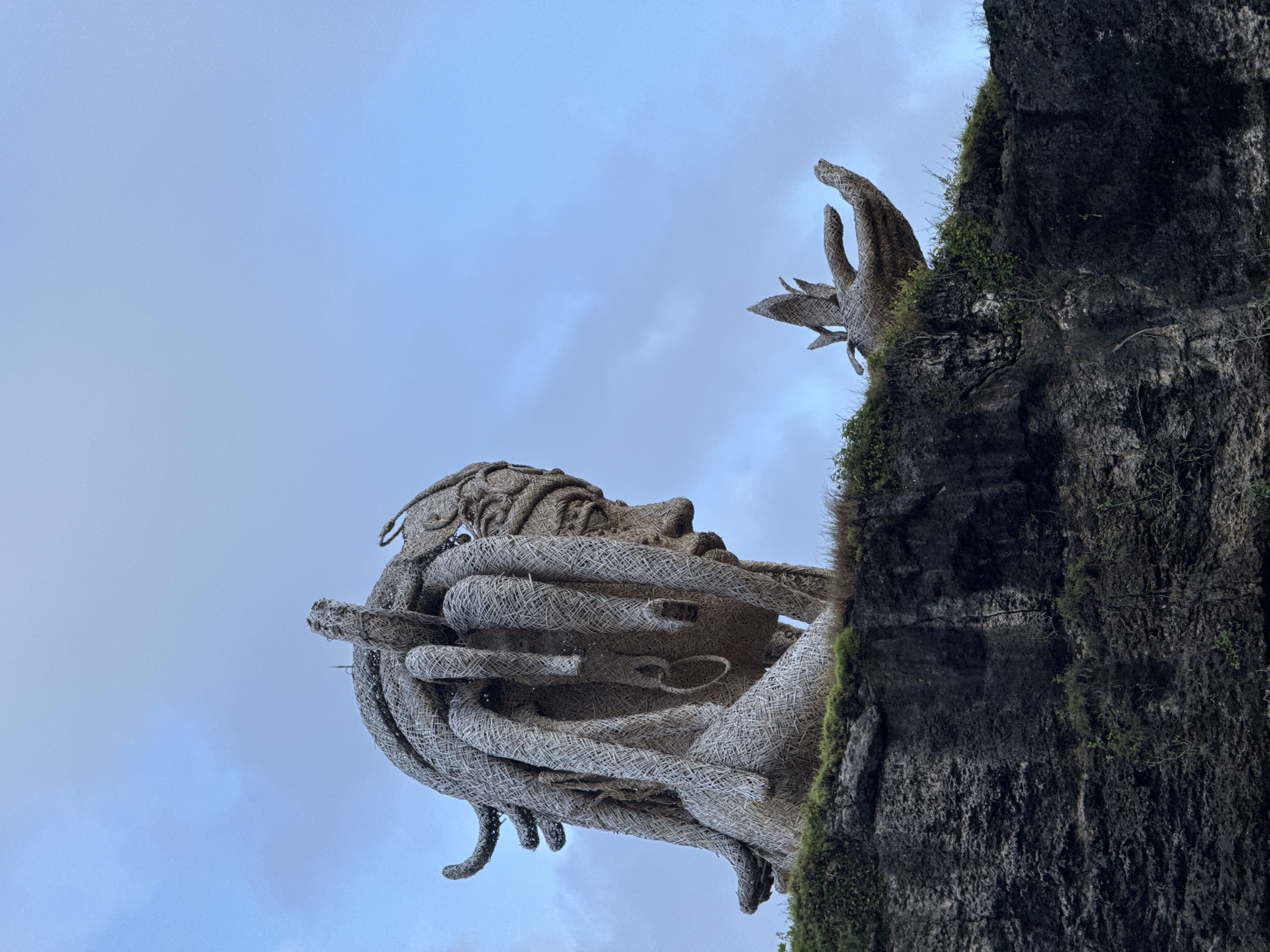 The photograph captures a low-angle view of a large woven sculpture depicting a human head and hand, situated on a moss-covered cliff. The sculpture appears to be made of a light-colored material resembling wicker or straw. The head is detailed, with discernible facial features and stylized hair. The hand extends upwards from the cliff face. The backdrop features a cloudy sky. The location is the island of Nusa Penida, Indonesia.