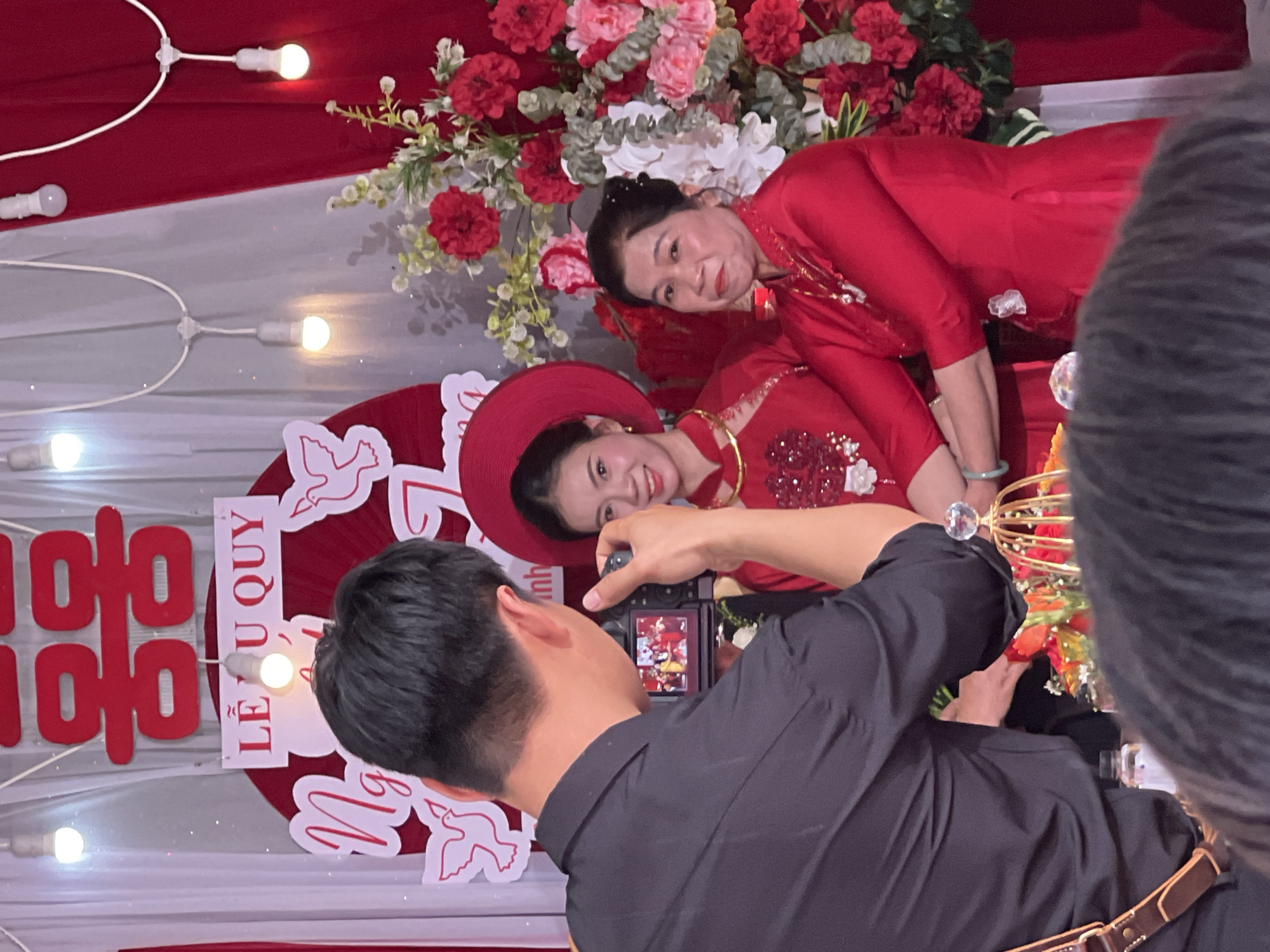 An indoor, festive scene in Tay Son Commune, Vietnam, captures a photographer taking a picture of two women during what appears to be a traditional Vietnamese engagement or wedding ceremony.
In the foreground, a man, presumably the photographer, is seen from behind, wearing a black shirt and holding a digital camera. He is focused on capturing the moment. His camera's screen displays the image he is composing.
Two women, dressed in elaborate red traditional Vietnamese attire, are posing for the camera. The woman closer to the photographer wears a wide-brimmed red hat and a gold necklace, smiling brightly. The woman behind her, also in a red costume with intricate detailing, has her arm resting gently on the first woman and is also smiling. Both women appear happy and relaxed.
The backdrop consists of draped red and white fabric, illuminated by warm, glowing string lights. A prominent, large floral arrangement featuring an array of red, pink, and white flowers is situated behind the women. To the left, a large red cutout of the "Double Happiness" (囍 - Song Hỷ) character, a common wedding symbol, is visible. Below it, a white rectangular sign clearly displays the Vietnamese text "LỄ VU QUY", which translates to "Bride's Departure Ceremony," confirming the celebratory nature of the event as a wedding or engagement. Additional decorative elements include white cutouts of doves and cursive script, partially visible as "Nguyệt Ánh" or similar. The overall ambiance is joyful and formal.