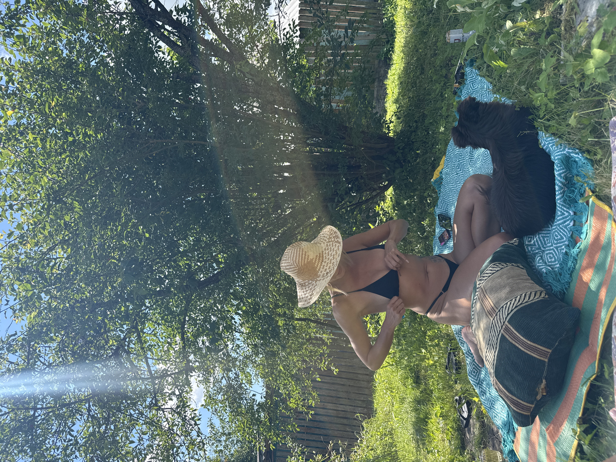 In the city of Krasnaya Presnya SNT, Russia, a blonde woman wearing a straw sun hat and a black bikini sits on a colorful blanket in a grassy, wooded area. Sunlight filters through the leaves of a large tree, casting dappled shadows. To her right, a small, dark-haired dog rests on the blanket. The woman is adjusting her bikini top. A wooden fence and other greenery are visible in the background, suggesting a backyard or garden setting.