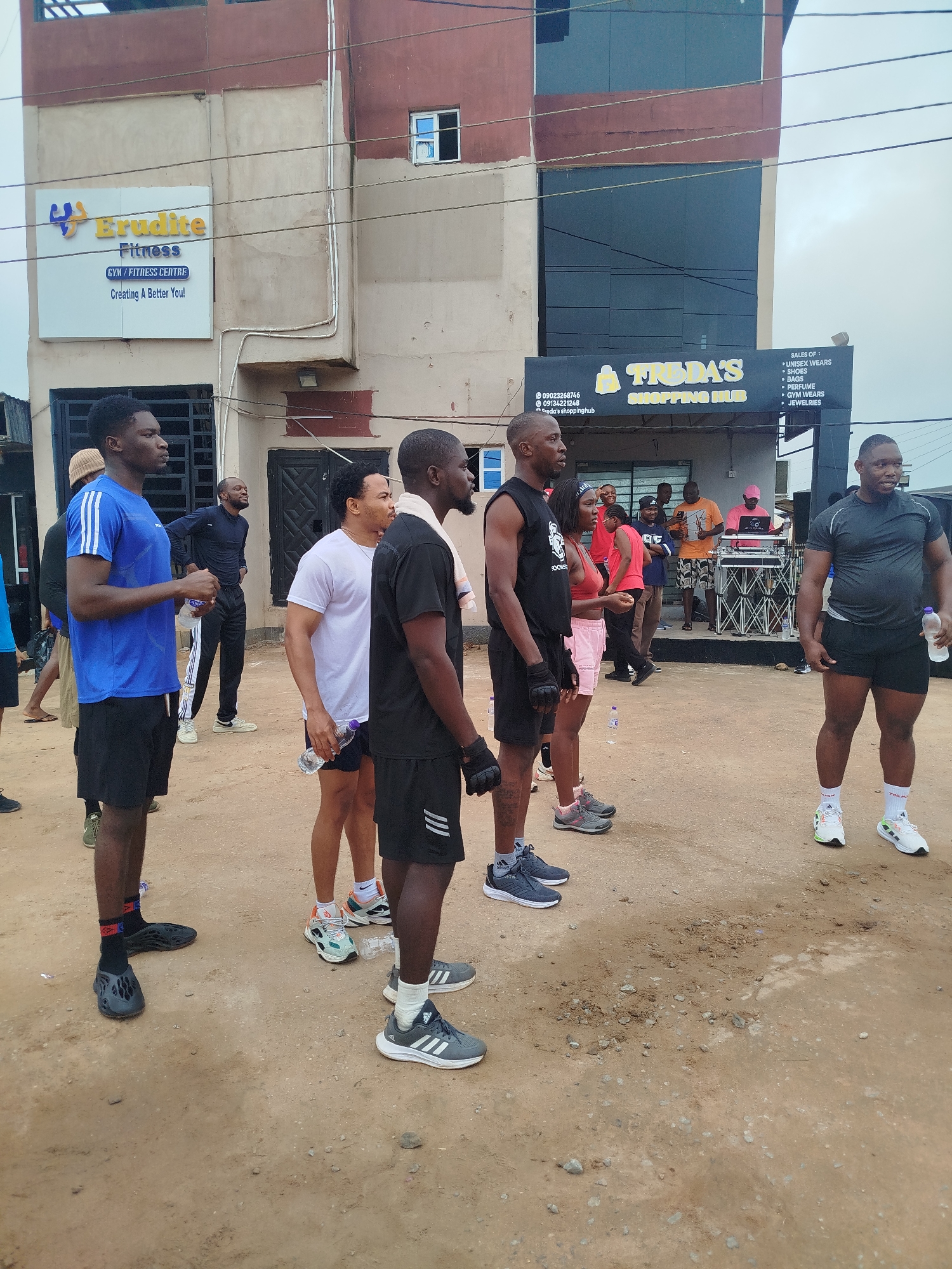 In Uselu, Nigeria, a group of people stands outside on a dirt ground in front of a building. The building has two signs, one for "Brudite Fitness" and another for "Tuda's Shopping Hub." A DJ setup with speakers is visible behind some of the people. The individuals are dressed in casual and athletic wear, with several holding water bottles. Some appear to be engaged in conversation or observing their surroundings. The overall scene suggests a public gathering, possibly related to fitness activities or an event at the shopping hub.