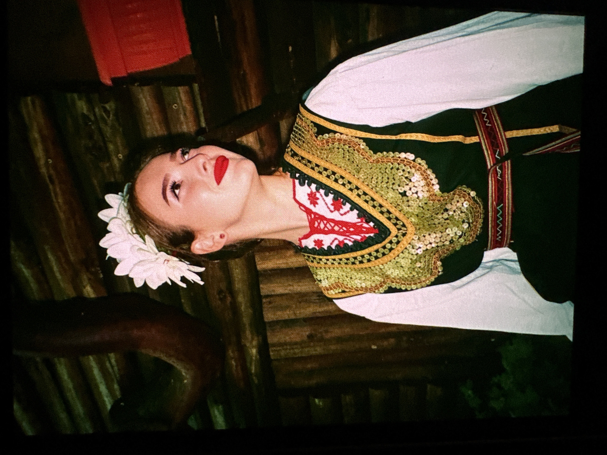 A young woman with bright red lipstick and a white flower crown on her head stands in front of a wooden wall. She is wearing a traditional Bulgarian folk costume, which includes a white blouse, a dark green vest adorned with gold trim and sequins, and a colorful embroidered belt. The background appears to be an outdoor setting, possibly a rustic building or a rural environment, with wooden planks and a glimpse of greenery. The woman is looking away from the camera, with a slight smile. The lighting suggests it may be evening or a dimly lit indoor space.