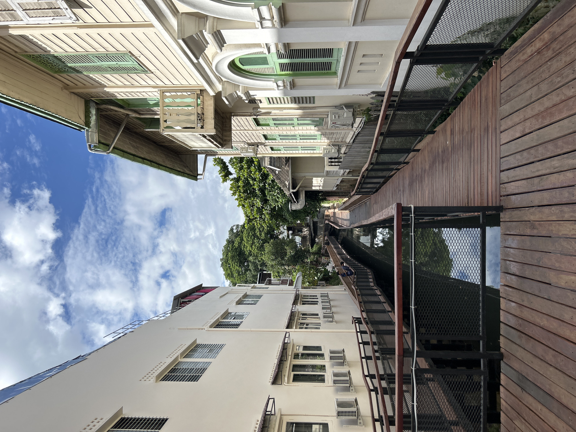 The image depicts a scenic walkway alongside a narrow canal in Chiang Mai City Municipality, Thailand. The walkway is constructed of weathered wooden planks and features dark metal railings with a mesh infill. On the left, a modern, cream-colored building with multiple windows and air conditioning units lines the canal. On the right, a traditional wooden building with green shutters and balconies adds a contrasting architectural element. Lush green trees are visible in the background, suggesting a verdant environment. The sky above is blue with scattered white clouds. Two individuals are seen walking on the wooden walkway in the middle distance, facing away from the camera. The canal water reflects the surrounding buildings and the sky, creating a serene atmosphere.