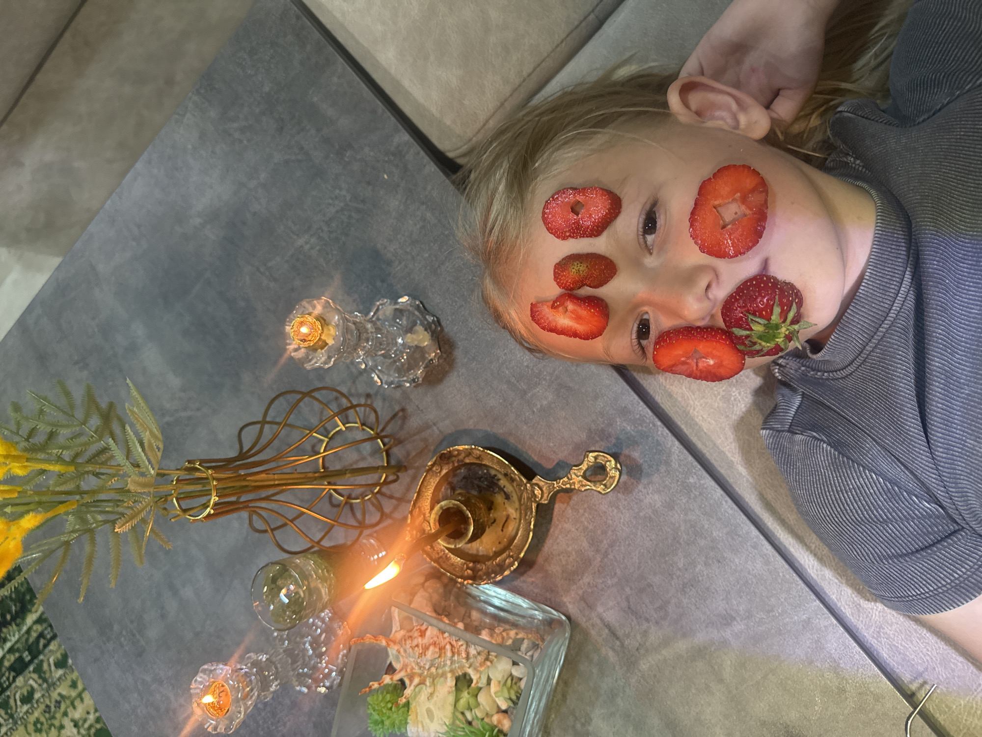 A close-up, angled shot shows a young child lying on a couch with pieces of strawberry arranged on their face as if in a spa treatment. One strawberry is whole and covers their upper lip, two halved strawberries are on their forehead and one on their cheek, with another strawberry with a hole in the center on their opposite cheek. The child is wearing a striped gray shirt and is surrounded by decorative items on a gray surface. To the left of the child, there is a glass vase with yellow flowers, several glass candle holders with lit candles, an ornate golden candlestick with a lit candle, and a glass container with shells and small plants. The environment appears to be an indoor living space. The location and city are unknown.