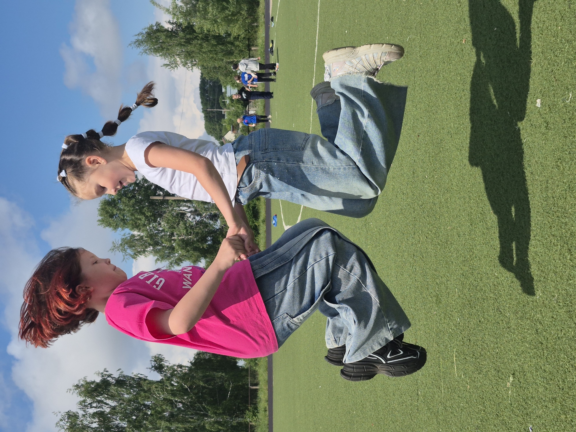 This rotated image features two young girls holding hands on an outdoor artificial turf field under a bright blue sky with scattered clouds. The perspective makes them appear suspended or floating horizontally, with their heads pointing towards the top of the frame.
The girl on the left has reddish-brown, chin-length hair and wears a bright pink short-sleeved t-shirt with "GLR WANG" partially visible on the front, baggy blue jeans, and black athletic shoes with white accents. She is looking towards the other girl.
The girl on the right has lighter brown hair styled into multiple small ponytails or braids secured with white hair ties. She wears a white short-sleeved t-shirt, baggy blue jeans, and light-colored athletic shoes. She is also looking towards her companion.
The girls are actively holding each other's hands, with their feet off the ground, suggesting they are mid-jump, balancing, or playing a game that involves lifting each other.
In the background, several other people, including adults and children, are visible further down the green field, which is bordered by trees. The presence of strong shadows on the artificial turf, including a prominent shadow of the photographer on the right, indicates bright sunlight, likely during midday or early afternoon. The scene depicts a lively outdoor gathering or recreational activity on a clear day.