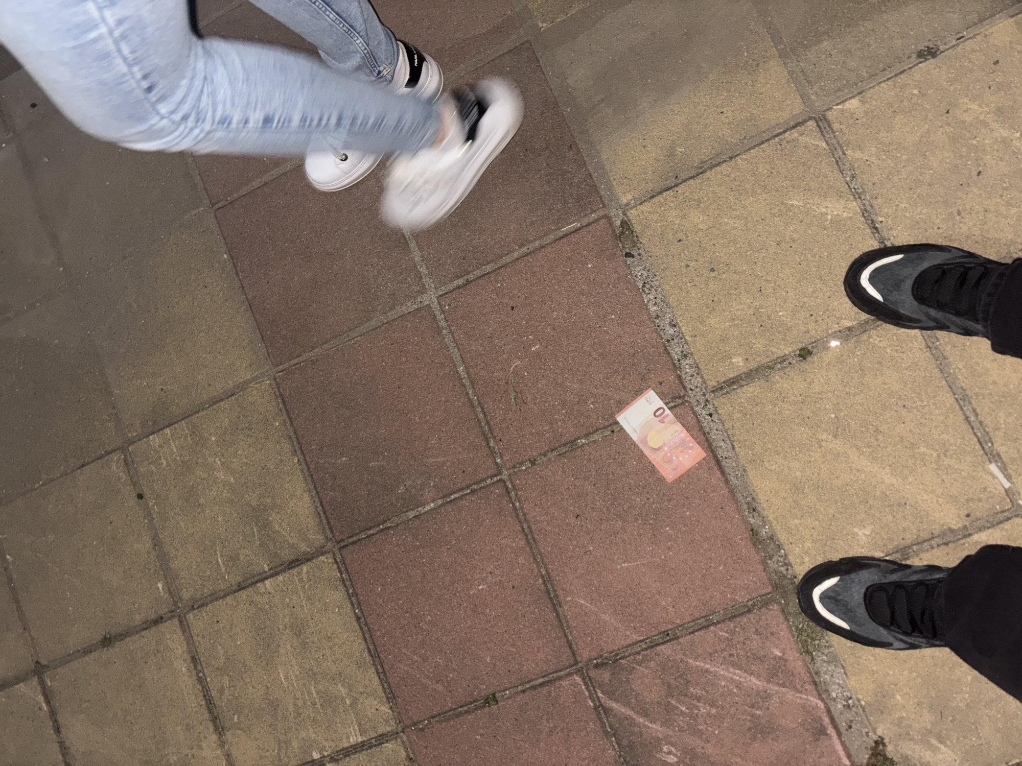 The image shows a paved ground with two people present, indicated by their shoes and lower legs. One person wears light blue jeans and white sneakers with black details, while the other wears black trousers and black sneakers with white accents. A 10 Euro banknote lies on the ground between them. The pavement consists of square tiles in alternating reddish-brown and yellow colors. The photograph seems to be taken outdoors, at night. The location is not identifiable as the city of Zlatograd, Bulgaria.