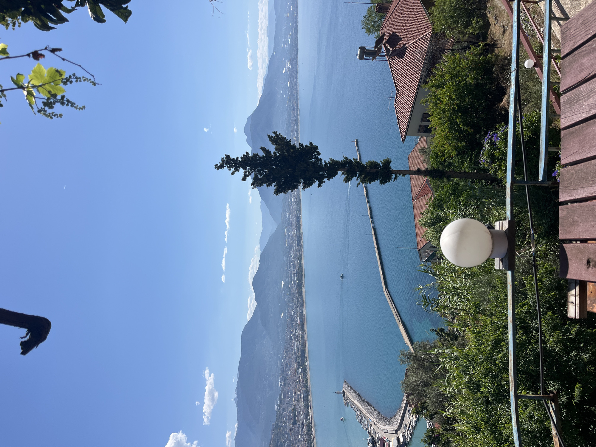 The image shows a view of Alanya, Turkey, from a high vantage point. A body of water, likely the Mediterranean Sea, fills the majority of the frame. A long breakwater stretches into the sea, enclosing a harbor on one end. The cityscape of Alanya is visible along the coastline, nestled against a backdrop of mountains. A tall, slender tree stands in the foreground. To the right, there are buildings with red-tiled roofs and lush green vegetation. A white spherical light fixture is also present. A portion of a wooden deck or walkway is visible on the right side. The sky is blue with scattered white clouds.
