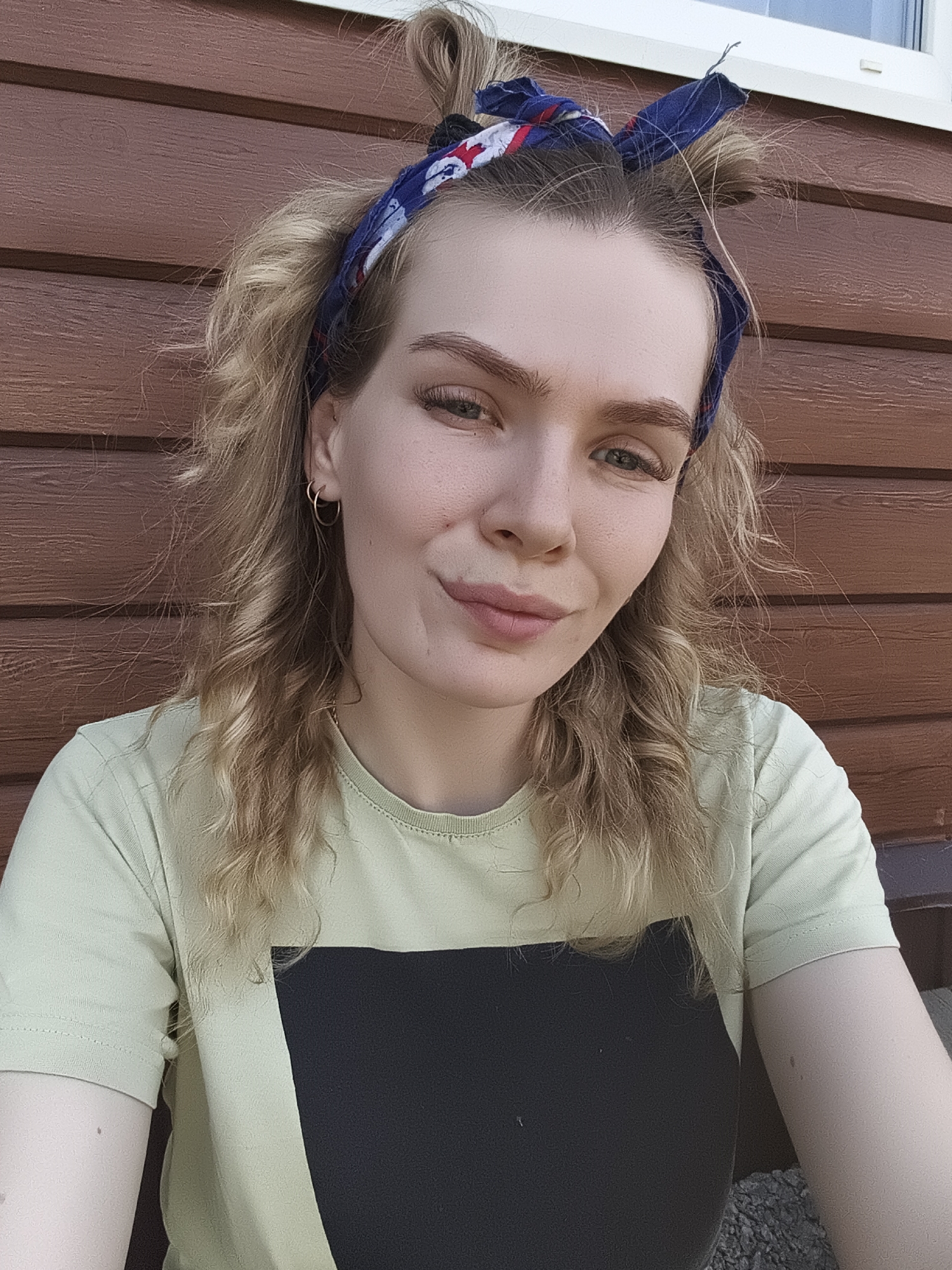 A young woman is positioned in front of a wooden wall, possibly the exterior of a building, with a window visible at the top right. The wall appears to be constructed of horizontal wood planks. She is wearing a light green t-shirt with a black square design on the front. Her hair is blonde, styled with loose curls and a blue and white bandana tied around her head with some hair pulled up. She is wearing small hoop earrings. Her facial expression includes a slight smile. The location cannot be verified as Русский Вожой, Russia, based on the provided image alone.