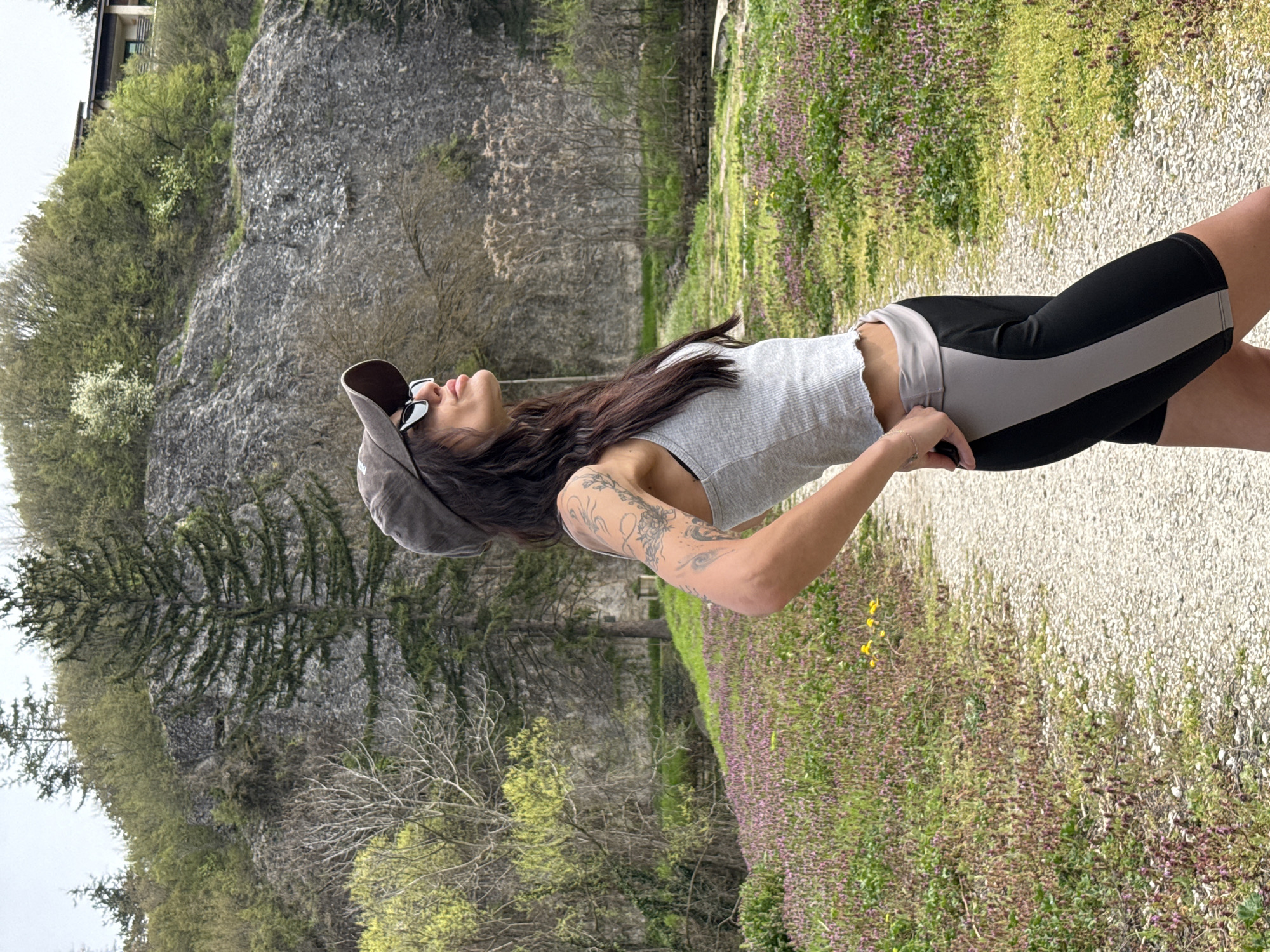 The image shows a woman standing on a gravel path next to a flower bed. The woman is wearing a grey tank top, black and grey shorts, a baseball cap, and sunglasses. She has long brown hair and tattoos on her arm. A large rock formation and trees are visible in the background. The city of Pleven, Bulgaria is not evident in the image.