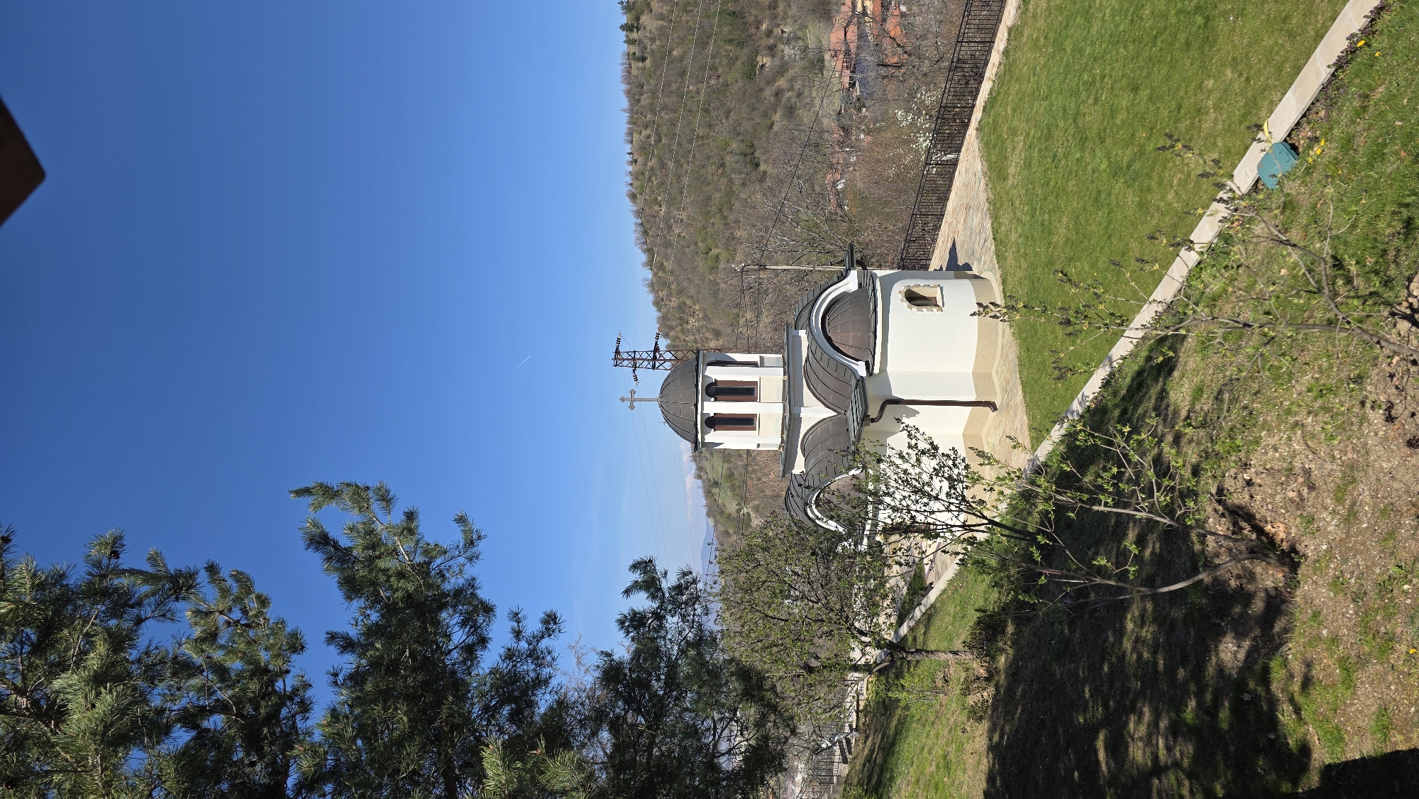 A medium-distance shot shows a small, white orthodox church with a dark gray domed roof topped with a cross, situated next to a grassy lawn. A mountain covered in trees is in the background, along with power lines, and a clear blue sky. In the foreground, to the left, is a pine tree, and other trees with budding leaves. The church appears to be located in a rural or semi-rural area. Based on the prompt, the location is in Elenovo, Bulgaria.