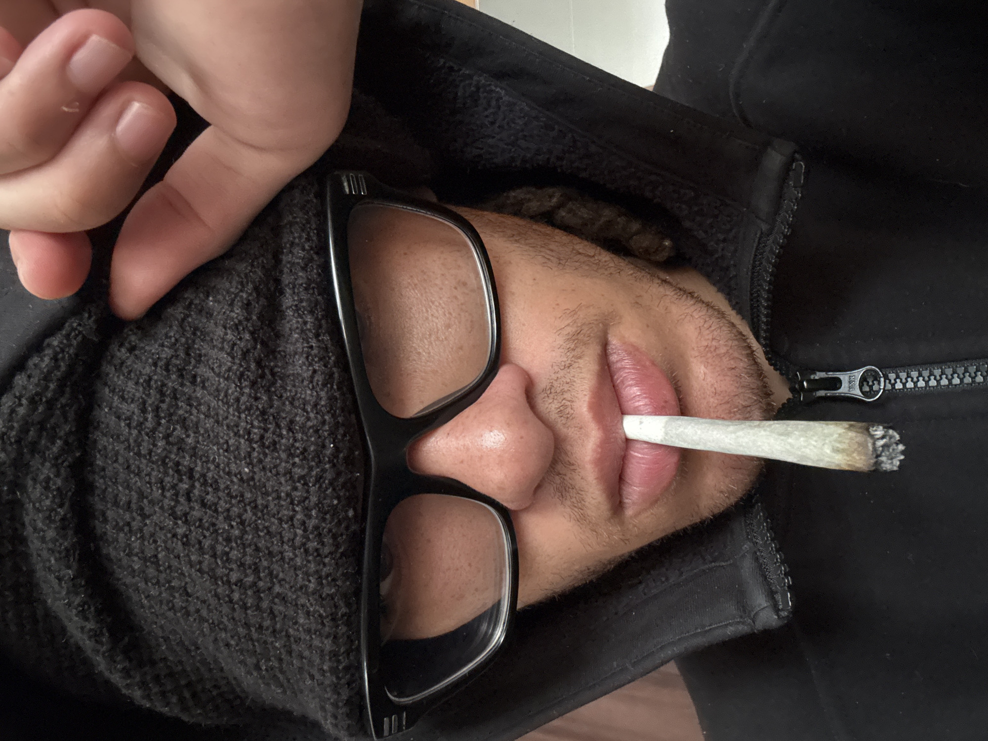 A close-up, rotated portrait captures a person from the chest up, seemingly lying down, with their head slightly turned. The individual is wearing black-rimmed glasses and a dark grey or black knitted beanie/hat that covers part of their head. A black jacket, possibly fleece-lined or a softshell, is worn, with a partially visible zipper that has "YKK" embossed on its pull tab.
The person has a lit, hand-rolled cigarette or joint held between their lips. Their facial features include visible stubble or short facial hair on the chin and upper lip, a distinct nose, and full lips. A hand with light skin tone is visible in the upper left corner of the frame, resting near the person's head, possibly their own hand or that of another individual. Dark, textured hair (possibly dreadlocks or thick curls) is glimpsed beneath the beanie. The background is indistinct, suggesting an indoor setting in Orsay, France. The overall impression is casual and relaxed.